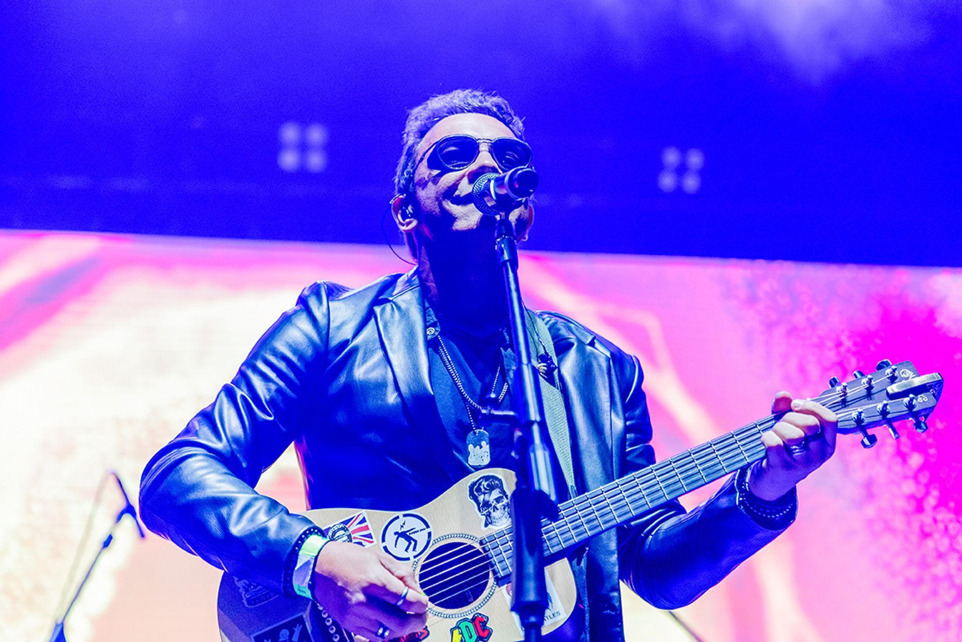 Um músico de jaqueta de couro toca um violão com adesivos enquanto se apresenta em um palco sob luzes fortes.
