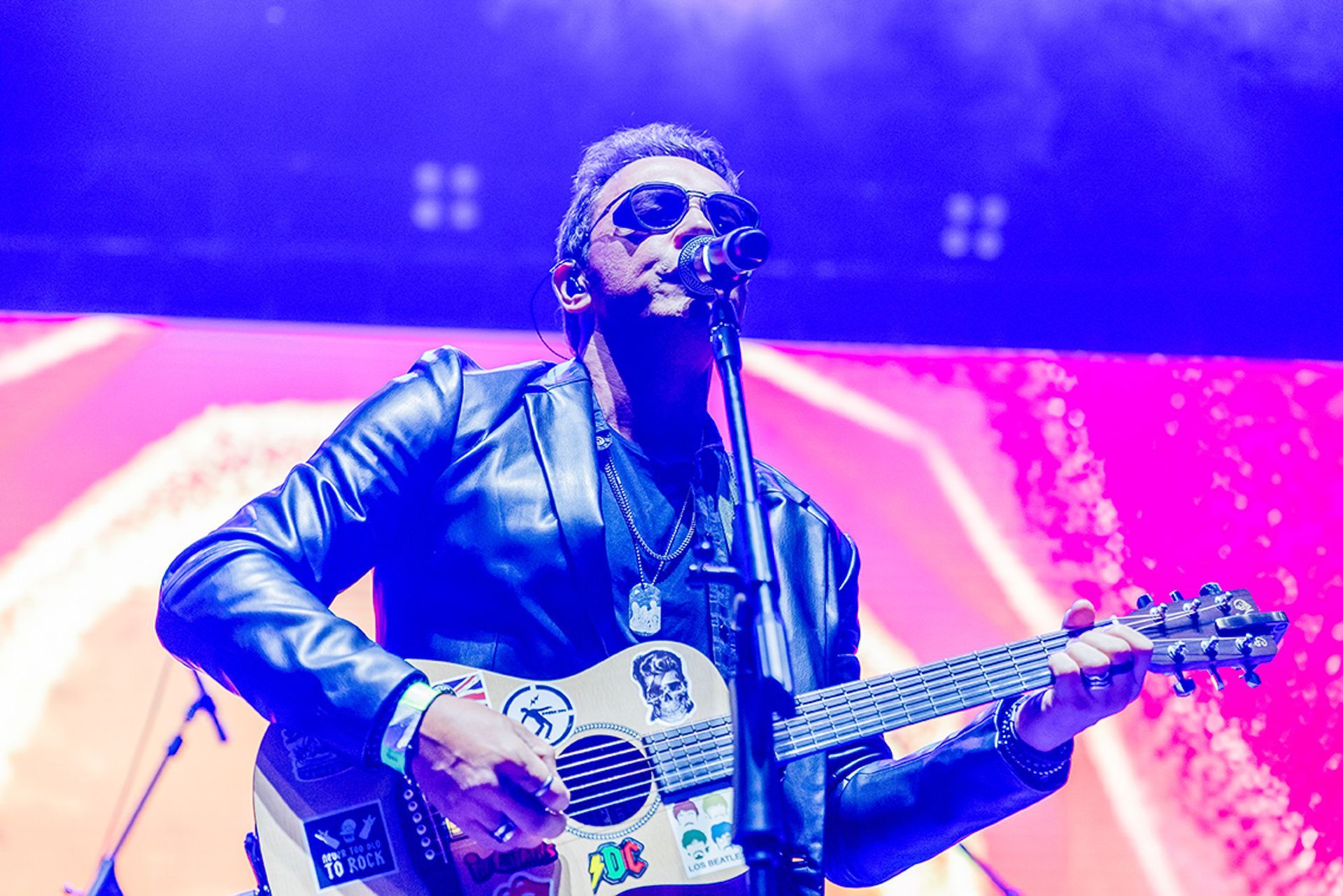 Um músico usando óculos escuros e jaqueta de couro toca um violão com adesivos enquanto se apresenta no palco.