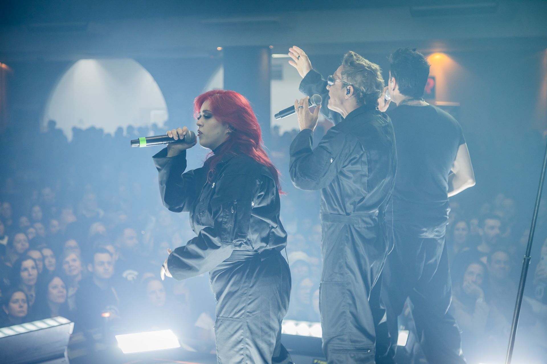 Três artistas vestindo macacões cinza iguais cantam em microfones para uma grande plateia em um show com pouca luz.