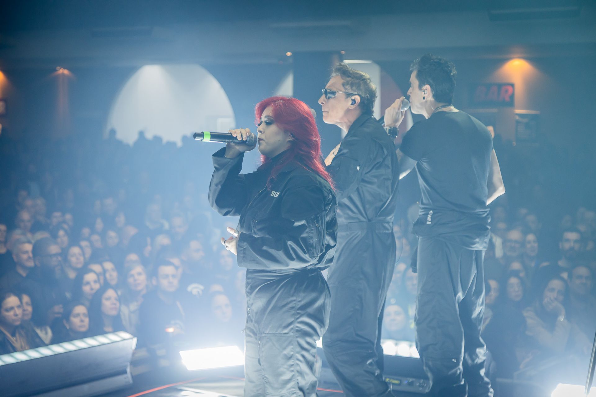 Três artistas em macacões escuros cantam em microfones num palco bem iluminado diante de uma grande plateia desfocada.