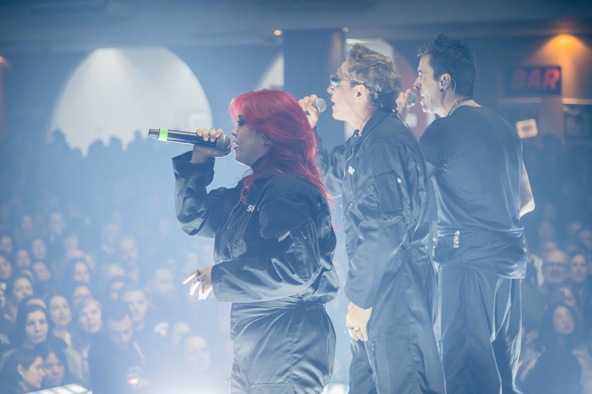 Um grupo musical se apresentando no palco com microfones diante de uma grande plateia com pouca luz.
