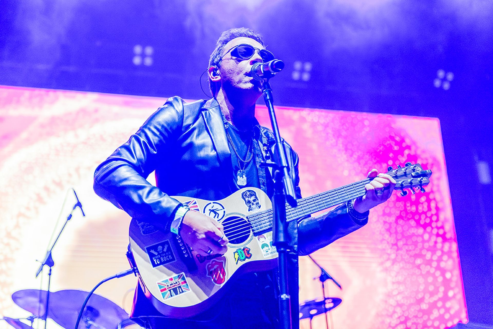 Um artista toca um violão coberto de adesivos enquanto canta em um microfone sob luzes azuis de palco.