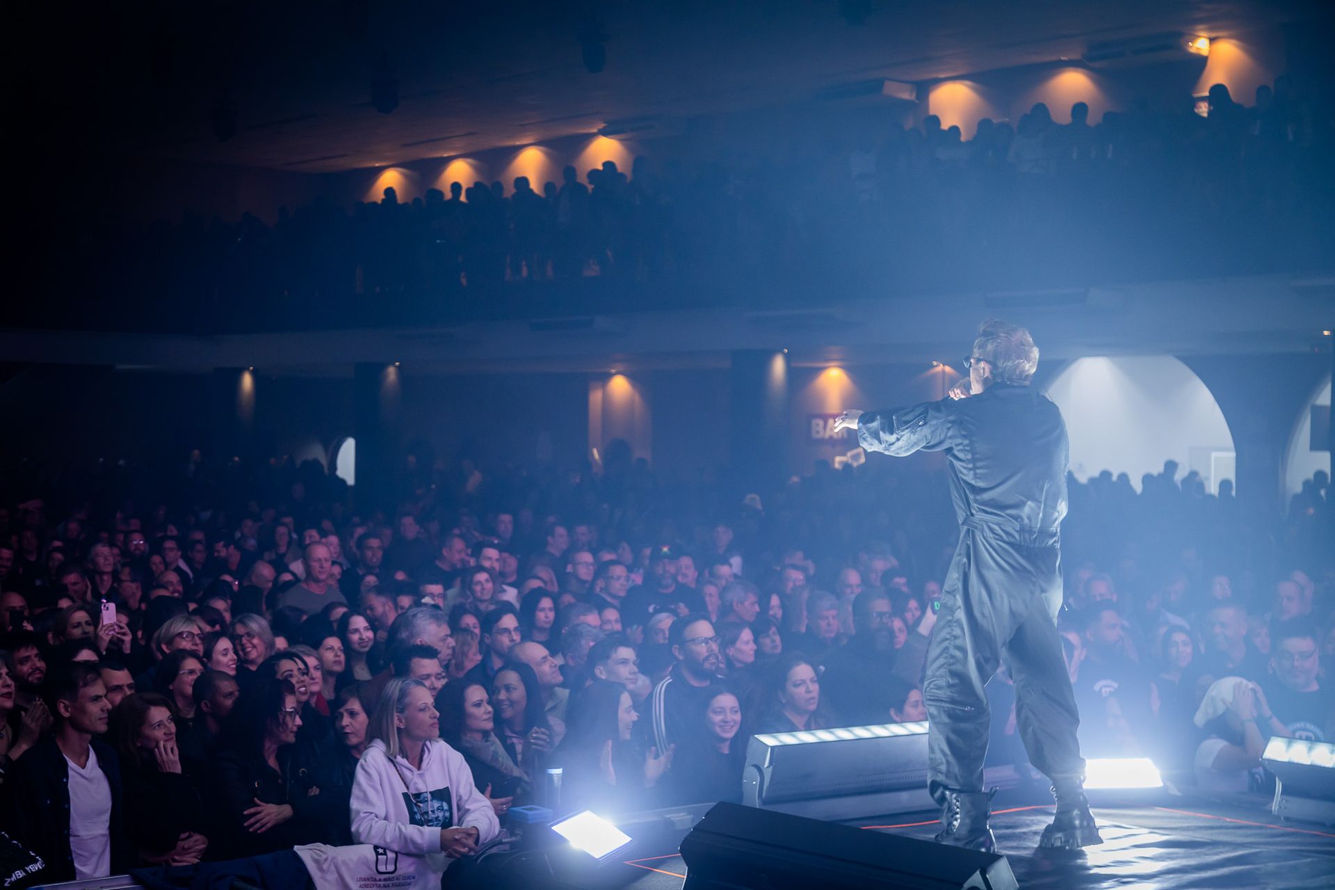 Um artista está em um palco pouco iluminado, gesticulando em direção a uma grande plateia sentada em um teatro.