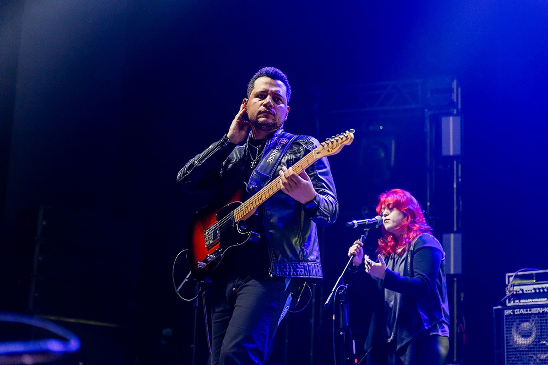 Um artista toca guitarra elétrica enquanto outra canta em um microfone em um palco iluminado com luz azul.