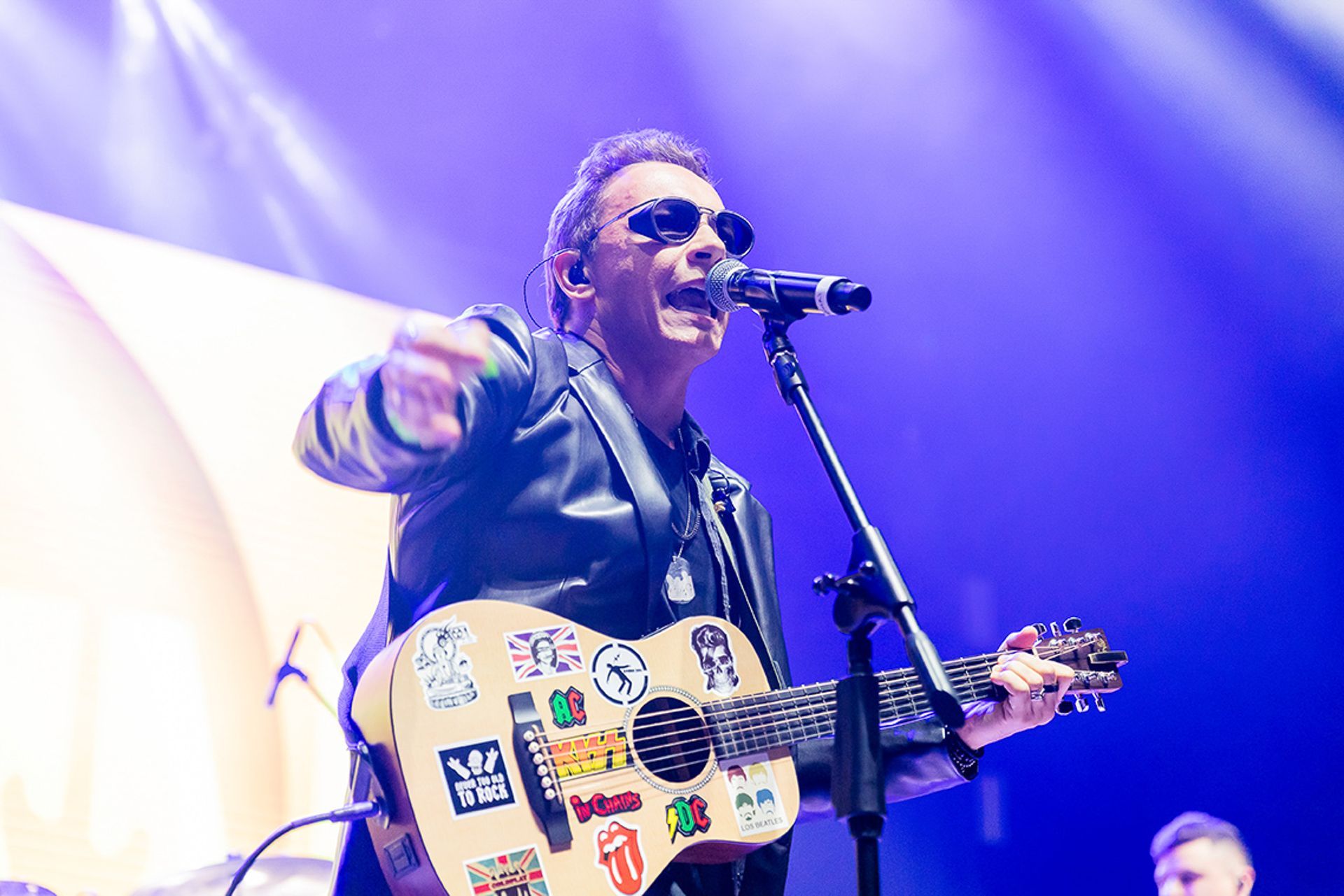 Um cantor de jaqueta de couro se apresenta no palco, tocando um violão coberto de adesivos em um microfone sob luz azul.