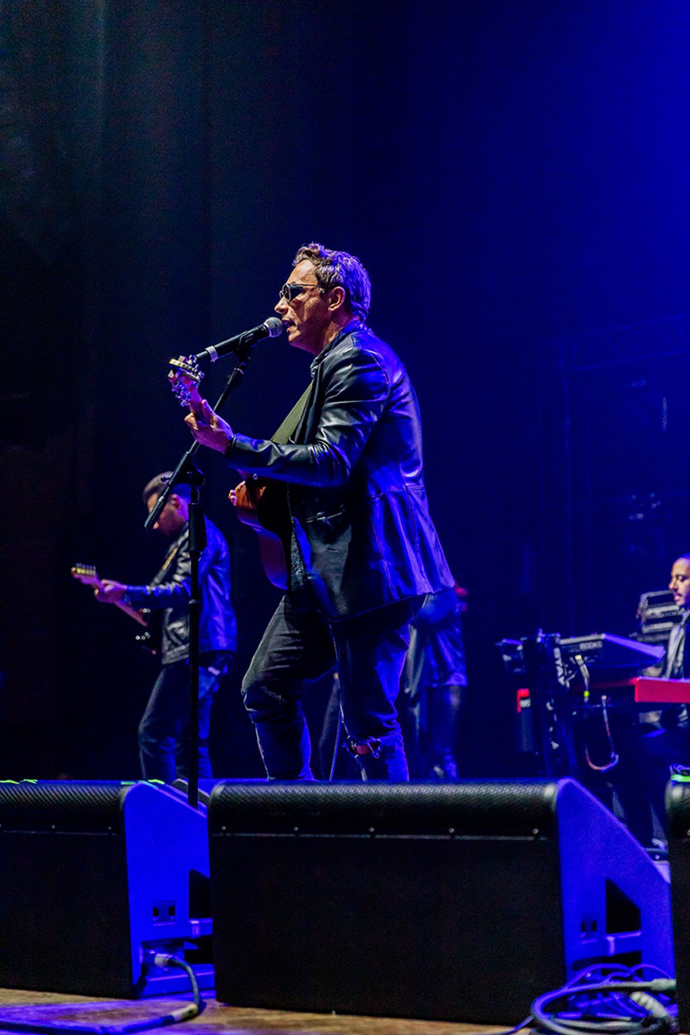 Um músico toca violão e canta em um microfone em um palco iluminado com luz azul, com seus companheiros de banda por perto.