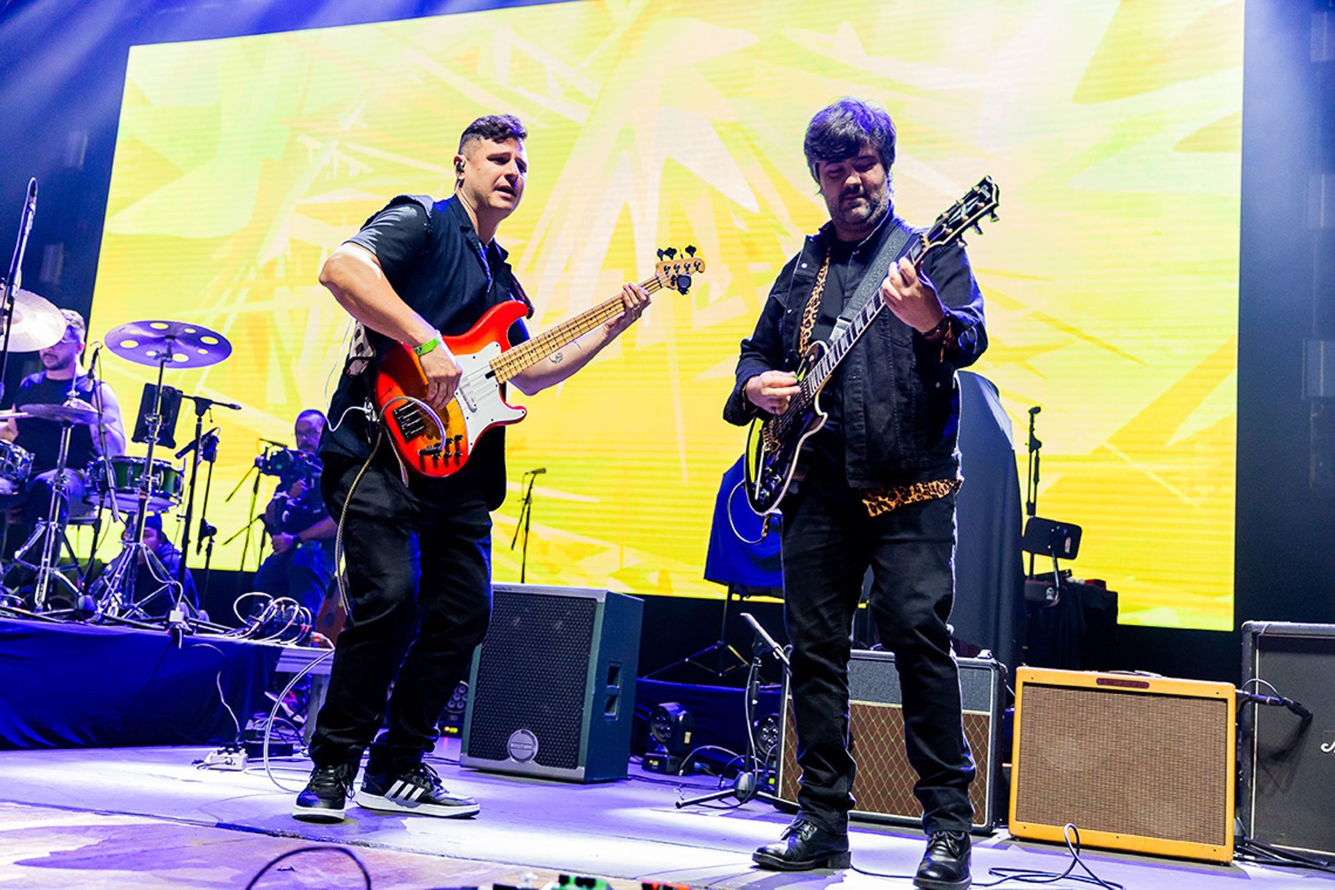 Dois músicos se apresentam no palco com guitarras elétricas, tendo como pano de fundo um amarelo