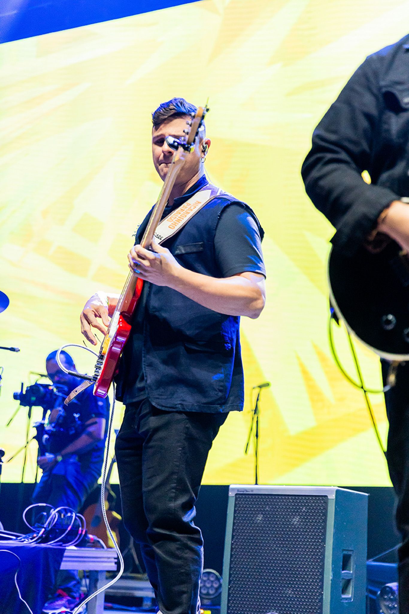 Um artista tocando uma guitarra elétrica vermelha no palco, em frente a um fundo amarelo brilhante.