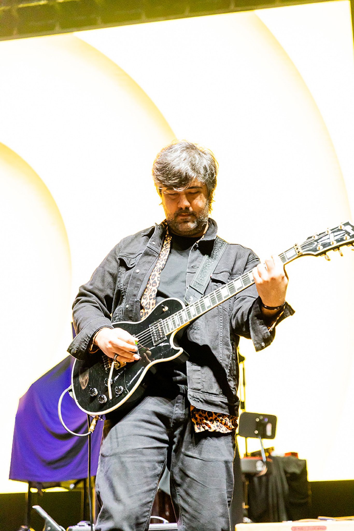 Um músico barbudo se apresenta no palco, tocando uma guitarra elétrica preta enquanto veste uma jaqueta escura.