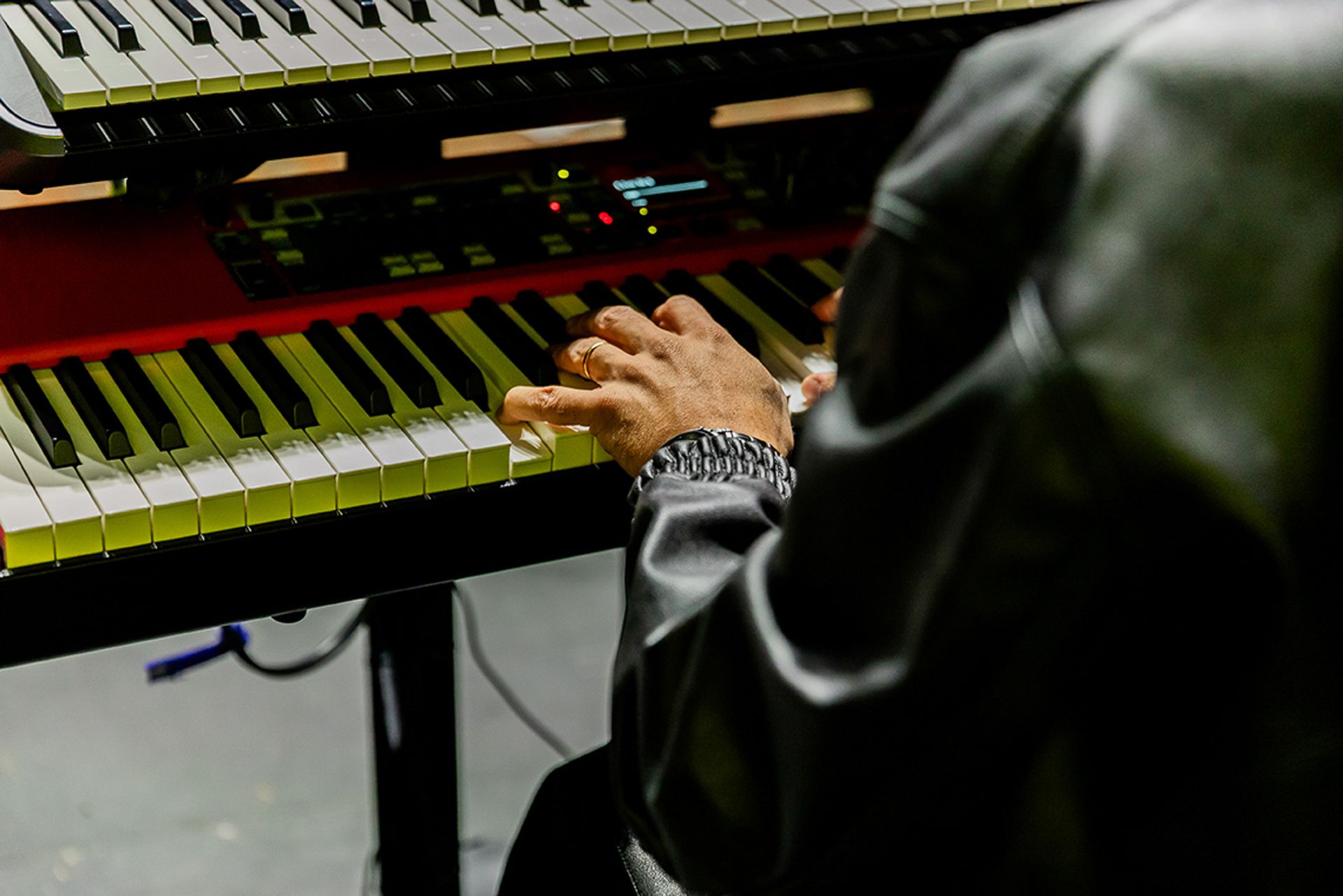 Um artista vestindo uma jaqueta de couro preta toca um teclado vermelho de dois níveis em um ambiente com pouca luz.