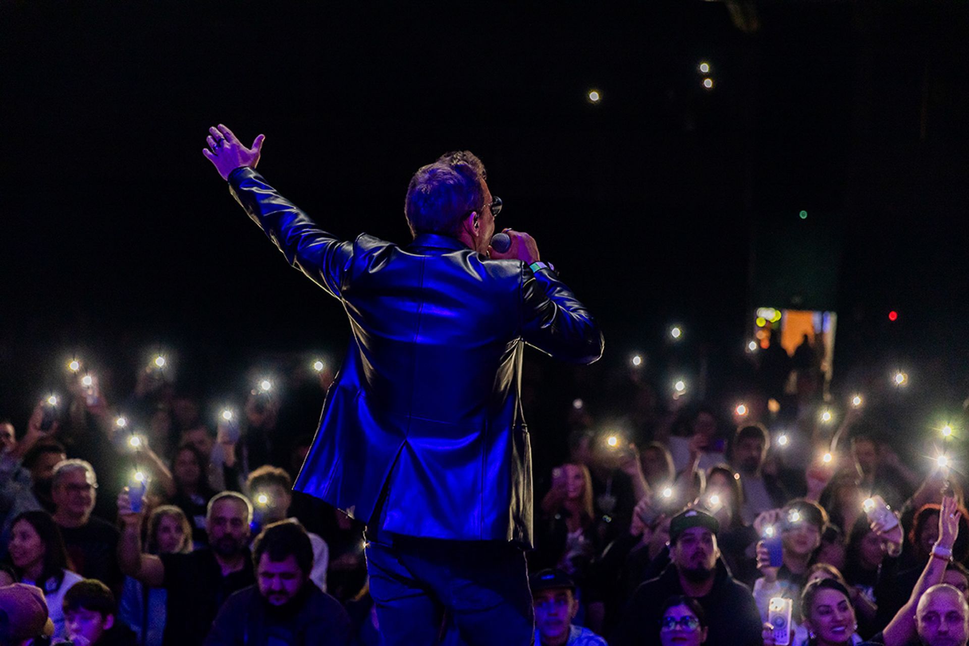 Um artista no palco, de costas para a câmera, gesticula em direção a uma plateia que segura as luzes acesas de seus celulares