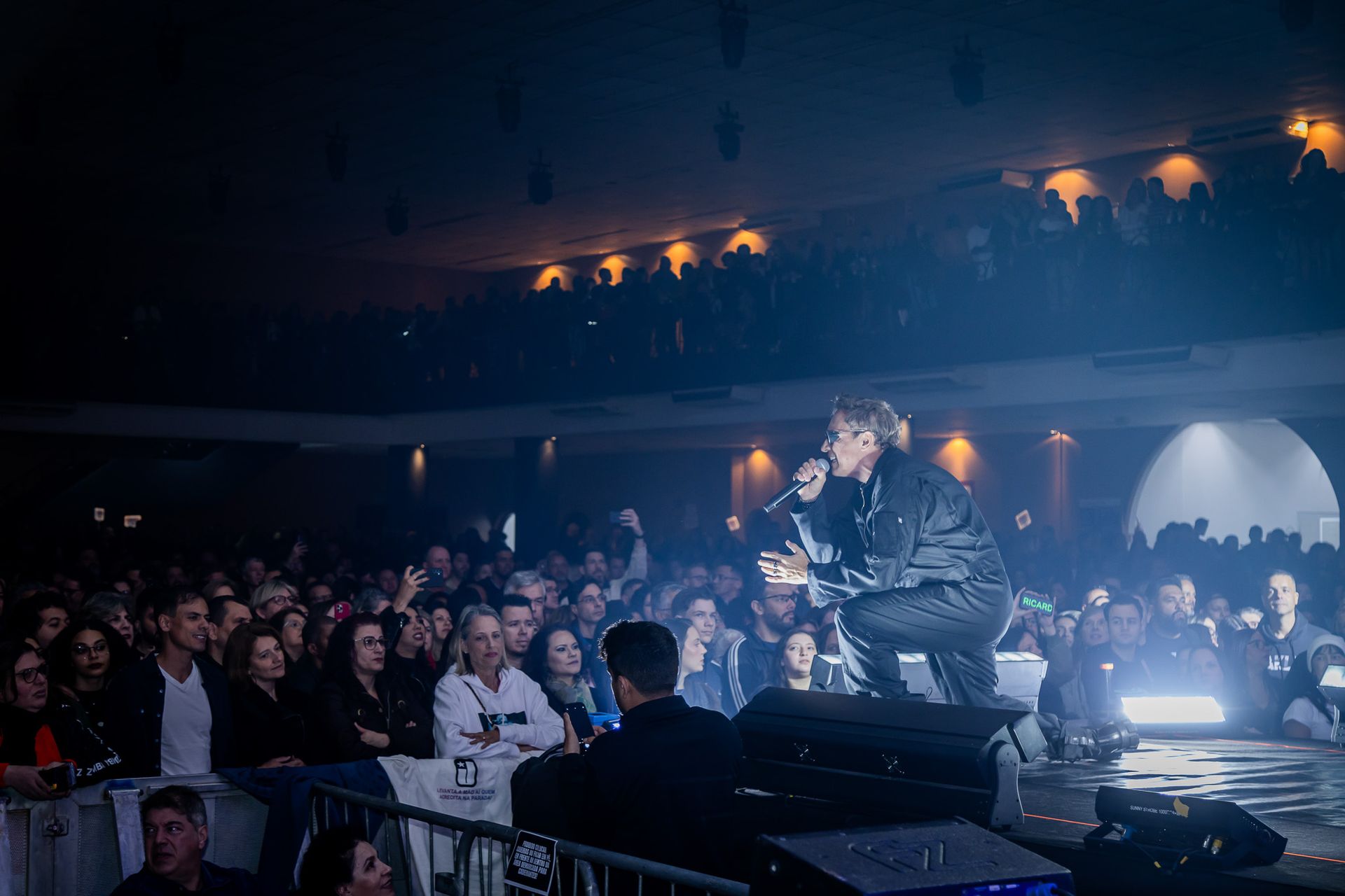 Um artista cantando ao microfone no palco diante de uma grande plateia com pouca luz em uma casa de shows.