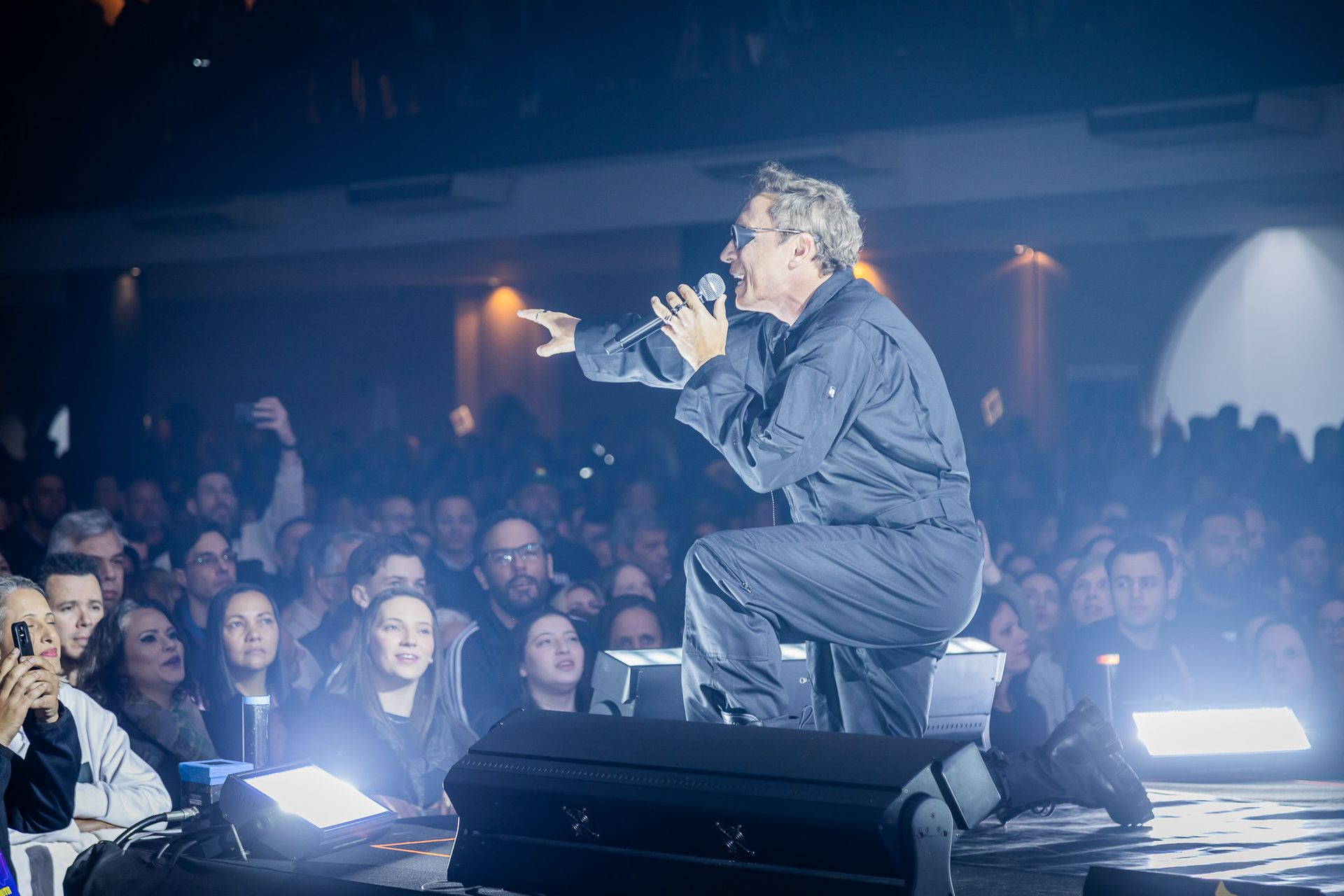 Um artista vestido com um macacão preto ajoelha-se no palco, cantando ao microfone diante de uma grande plateia em um show.
