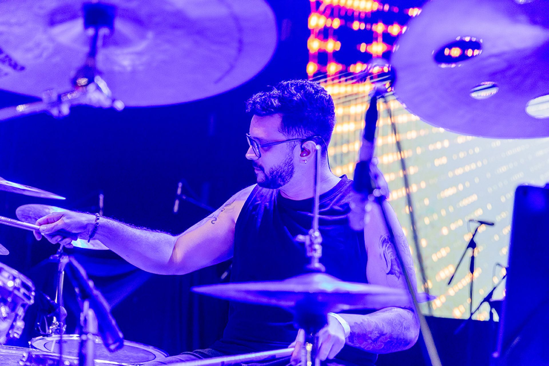 Um baterista de regata preta toca bateria no palco sob uma iluminação roxa brilhante, cercado por pratos.