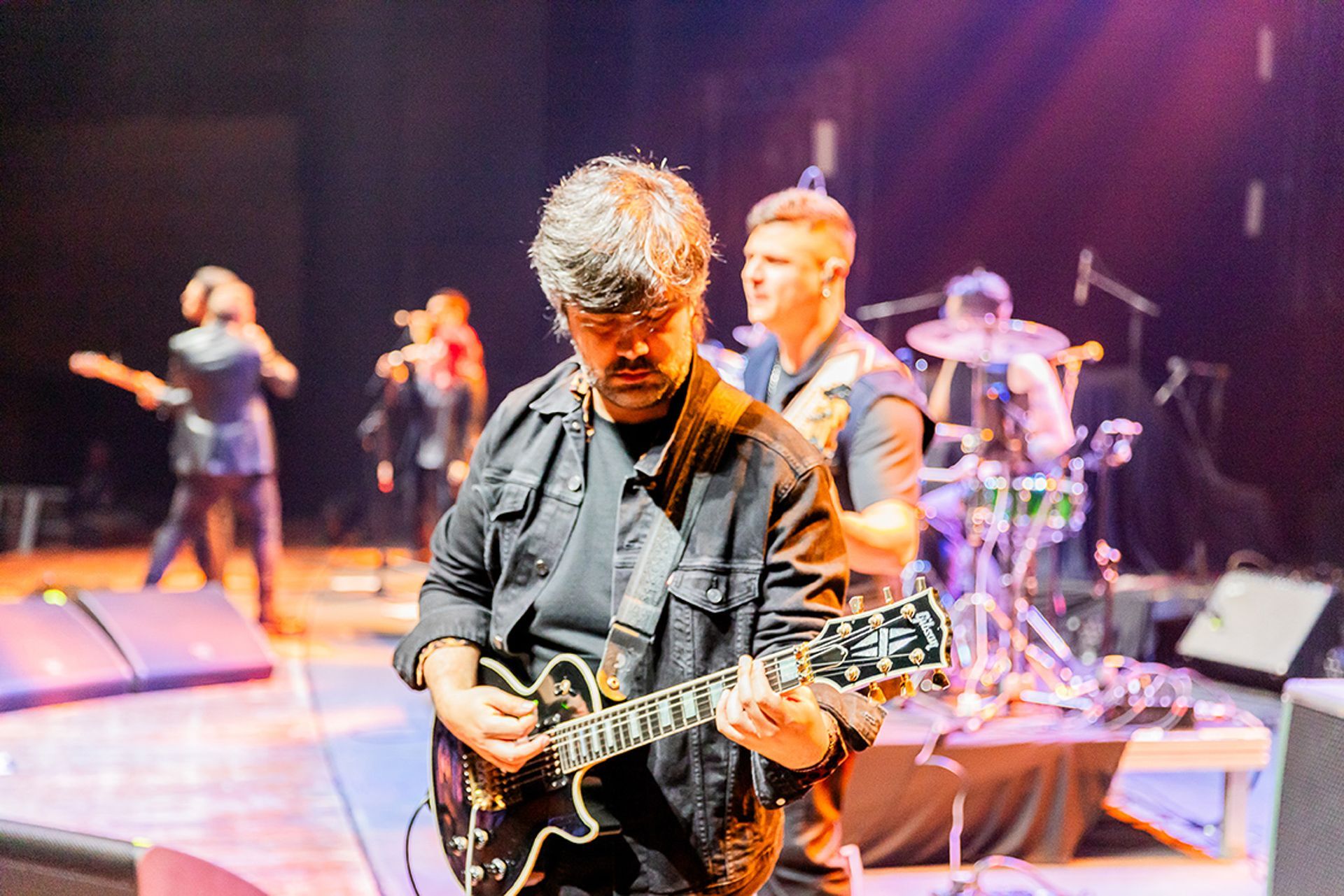 Um músico toca guitarra elétrica em um palco com uma banda, sob iluminação forte e com fundo escuro.