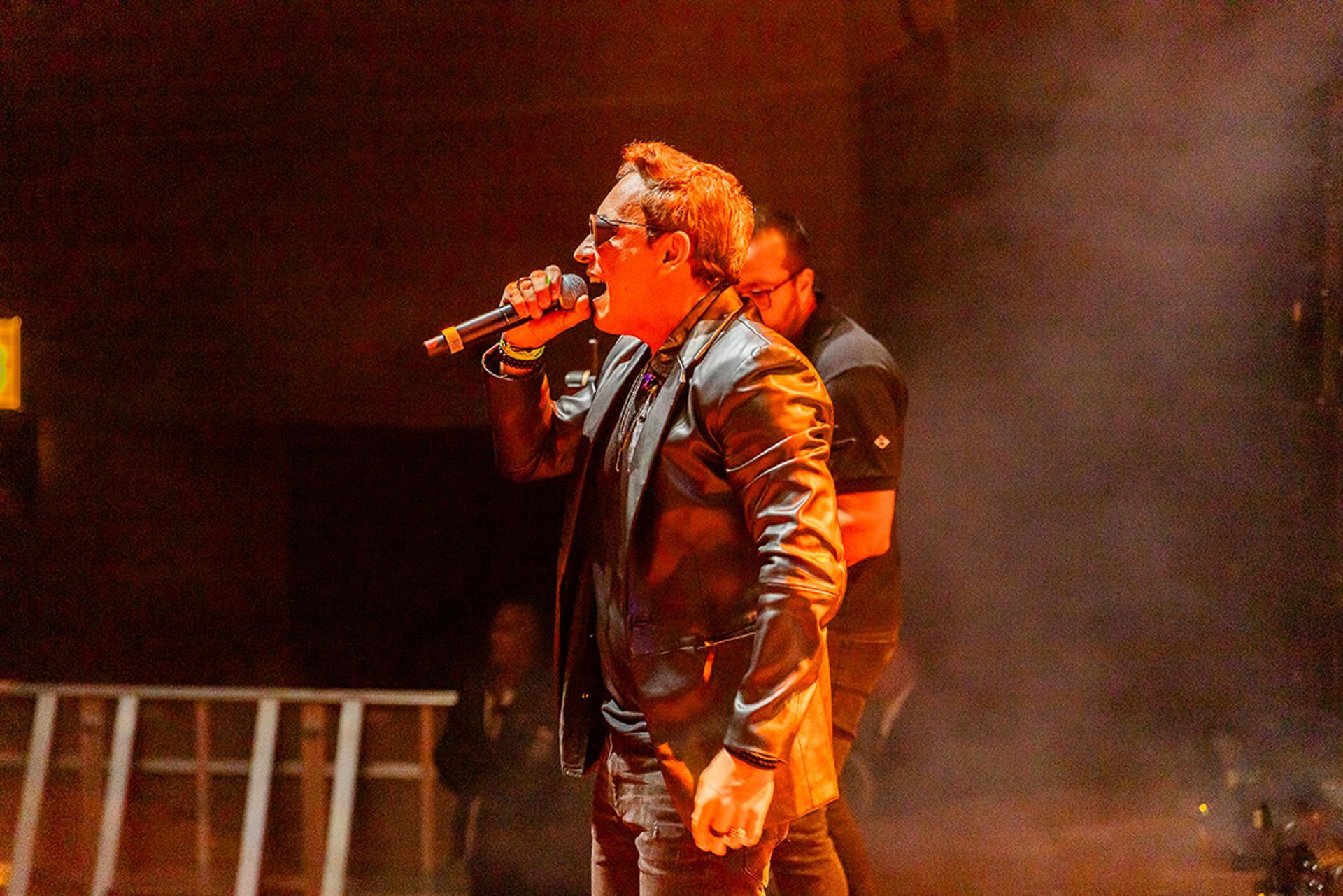 Um cantor com jaqueta metálica se apresenta no palco com um microfone sob iluminação quente.