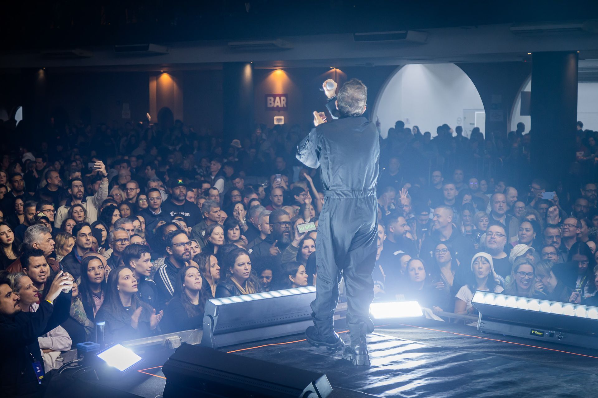 Um artista vestido de escuro está em um palco bem iluminado, de frente para uma grande plateia em uma sala de concertos.