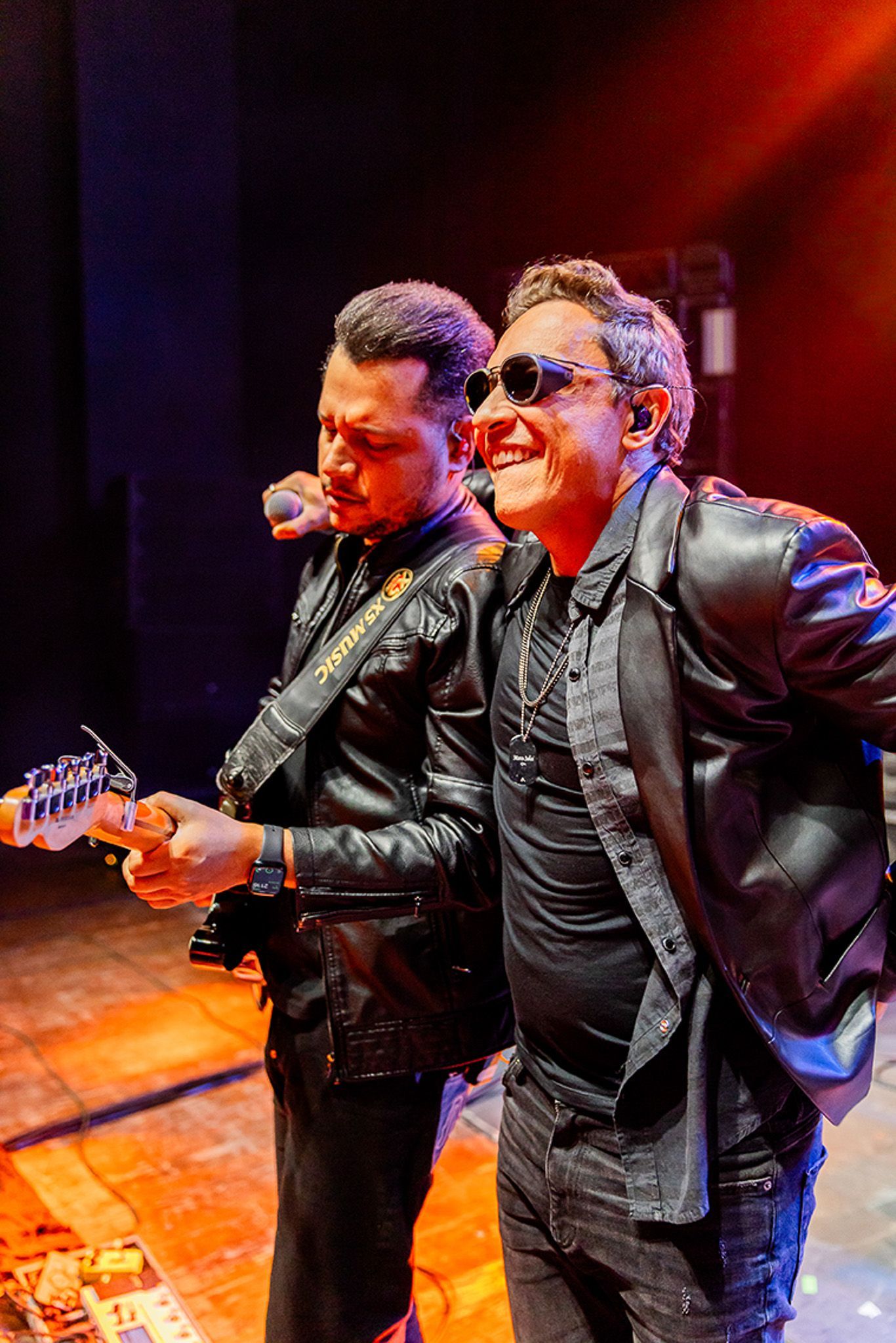 Dois músicos com jaquetas de couro pretas se apresentam em um palco um tocando guitarra elétrica e o outro cantando.