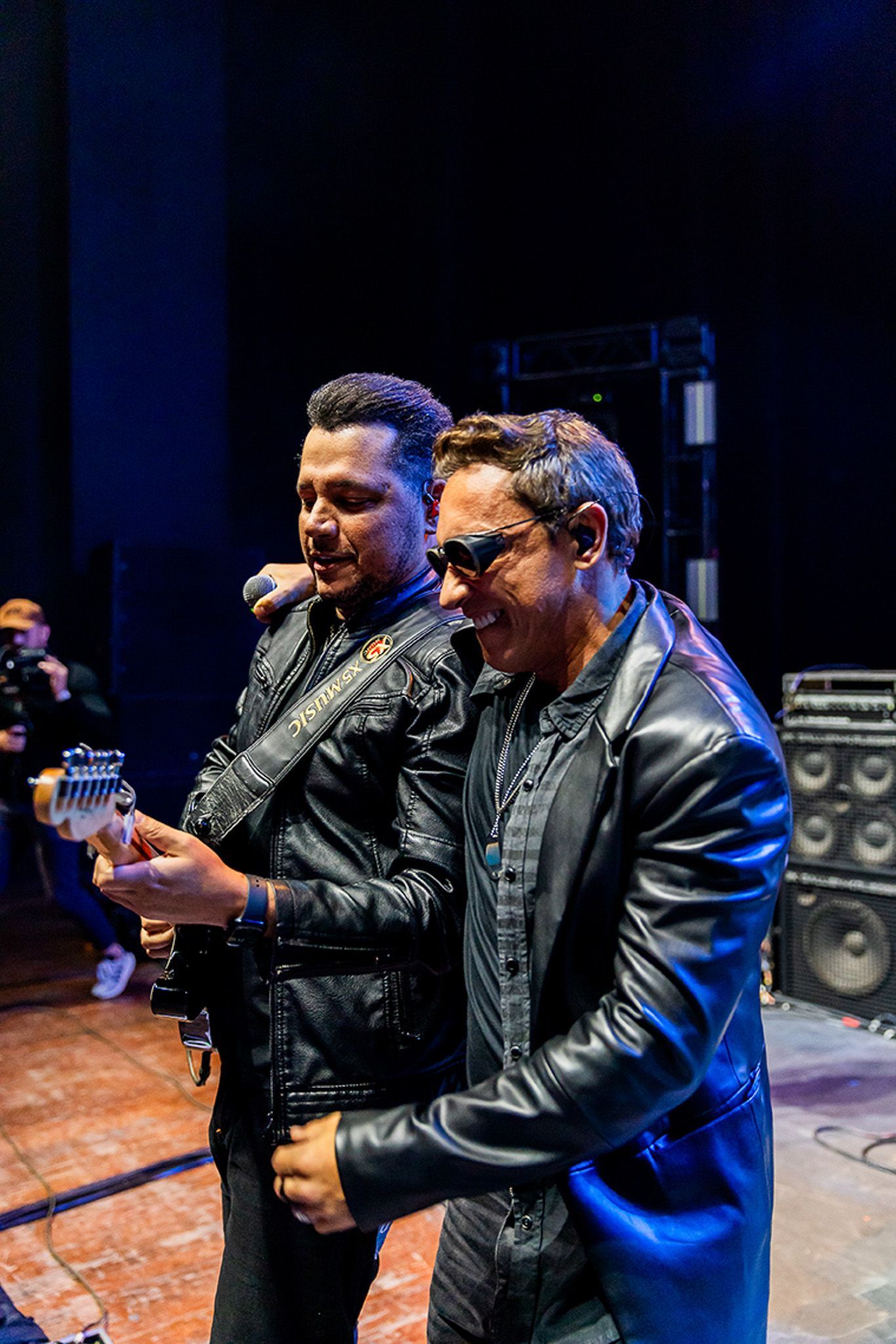 Dois músicos com jaquetas de couro pretas se apresentam, um tocando guitarra elétrica enquanto o outro sorri ao seu lado.