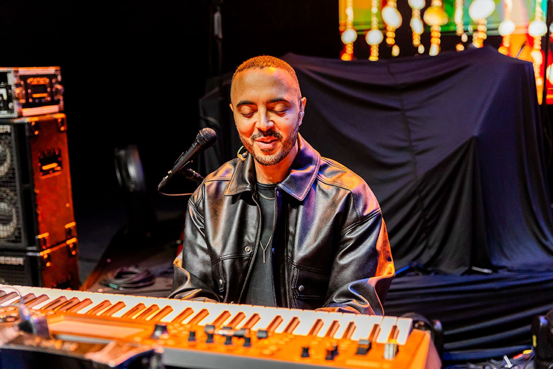 Um artista vestindo uma jaqueta de couro toca teclado no palco, sorrindo enquanto se apresenta em frente a um microfone.