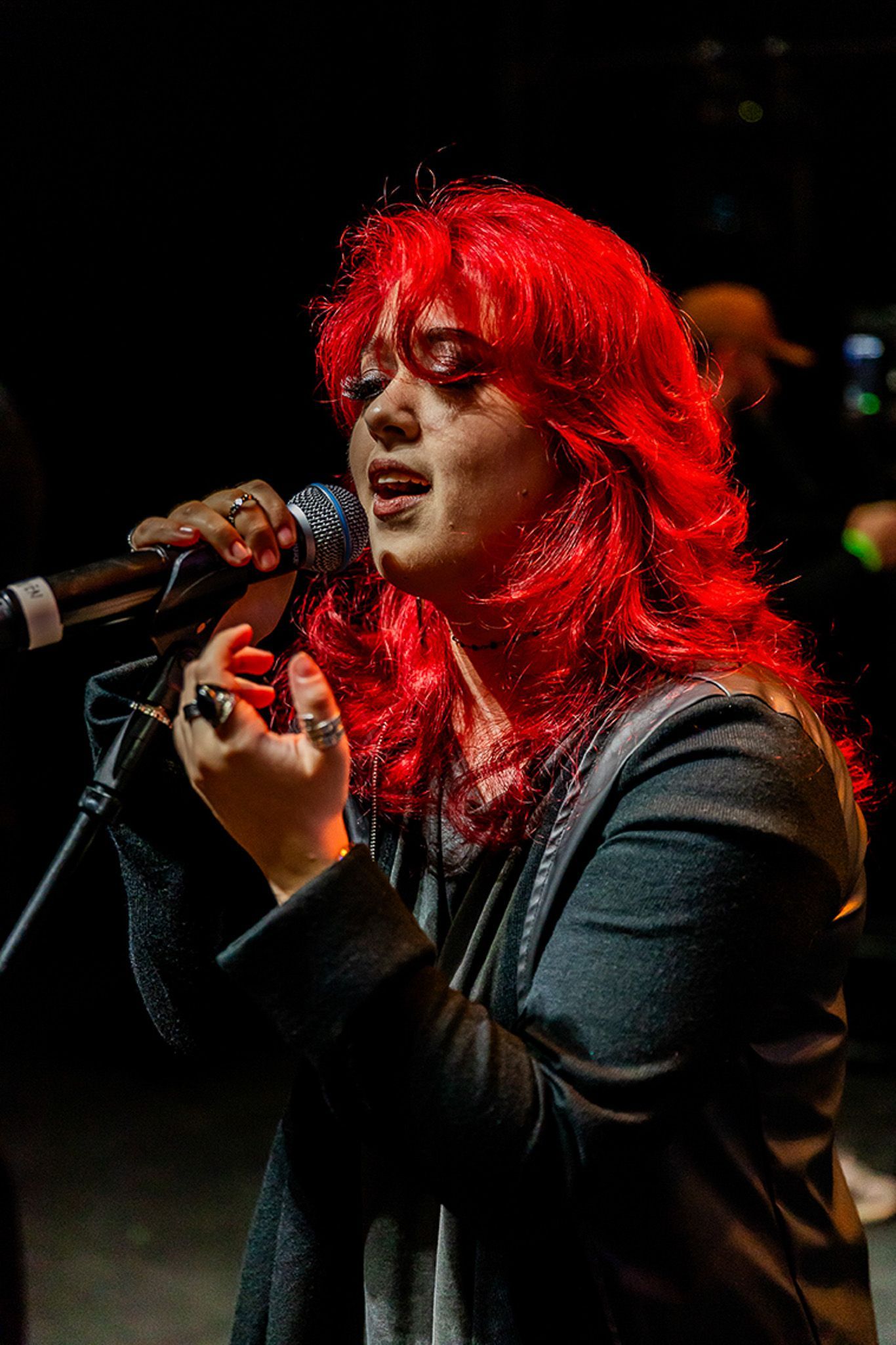 Uma cantora de cabelos ruivos vibrantes se apresenta ao microfone em um palco escuro.