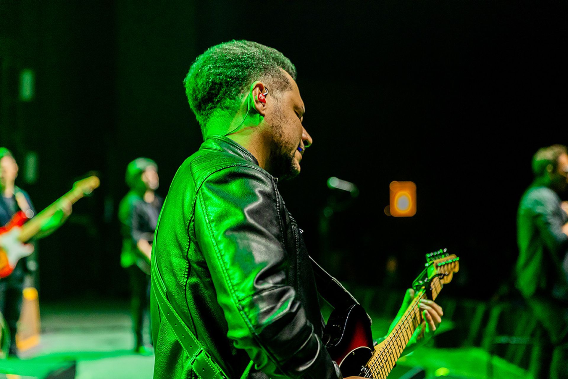 Um músico com jaqueta de couro preta toca guitarra elétrica no palco sob luzes verdes de concerto.