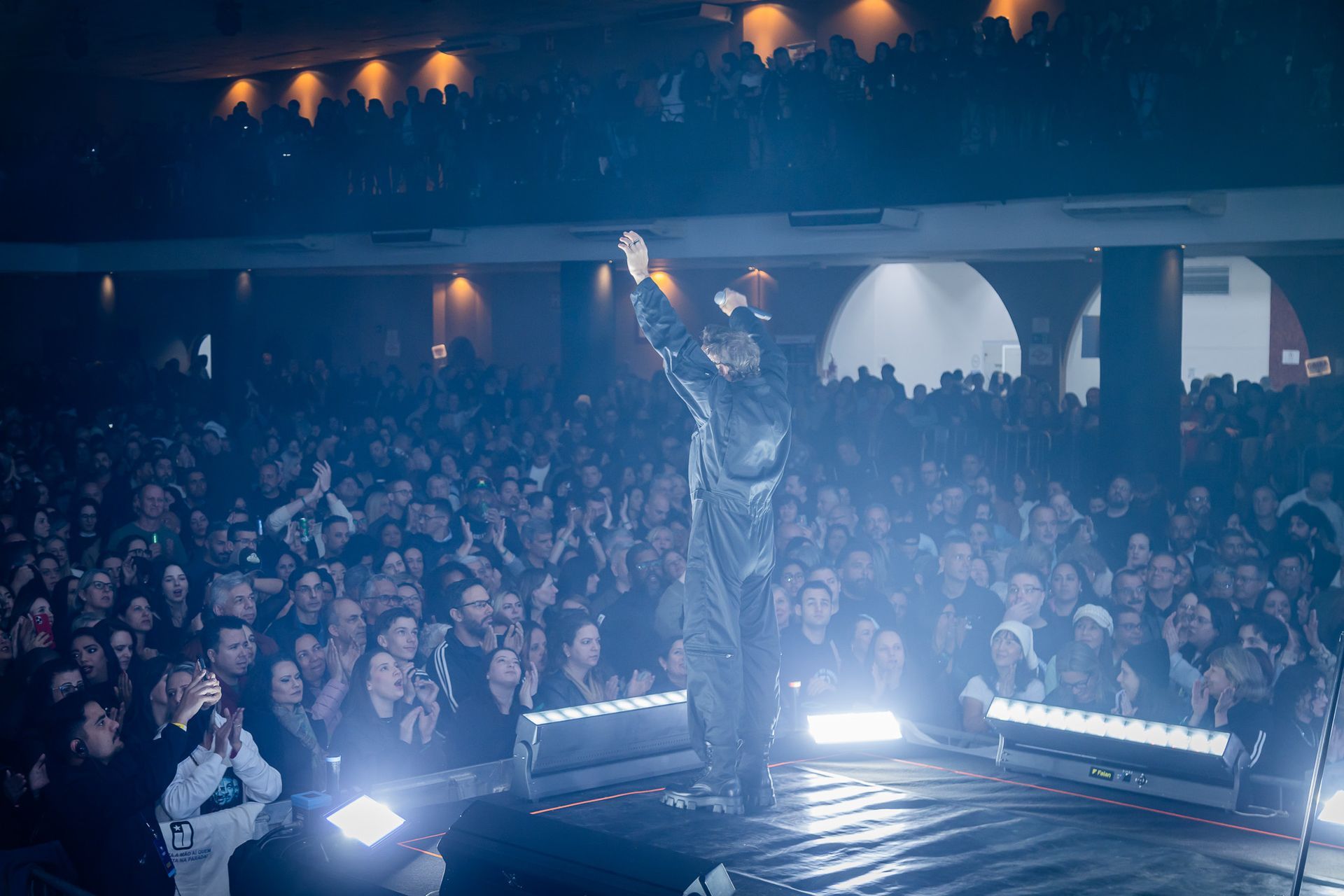 Um artista está em um palco pouco iluminado, com o braço erguido diante de uma grande plateia que o aplaude.