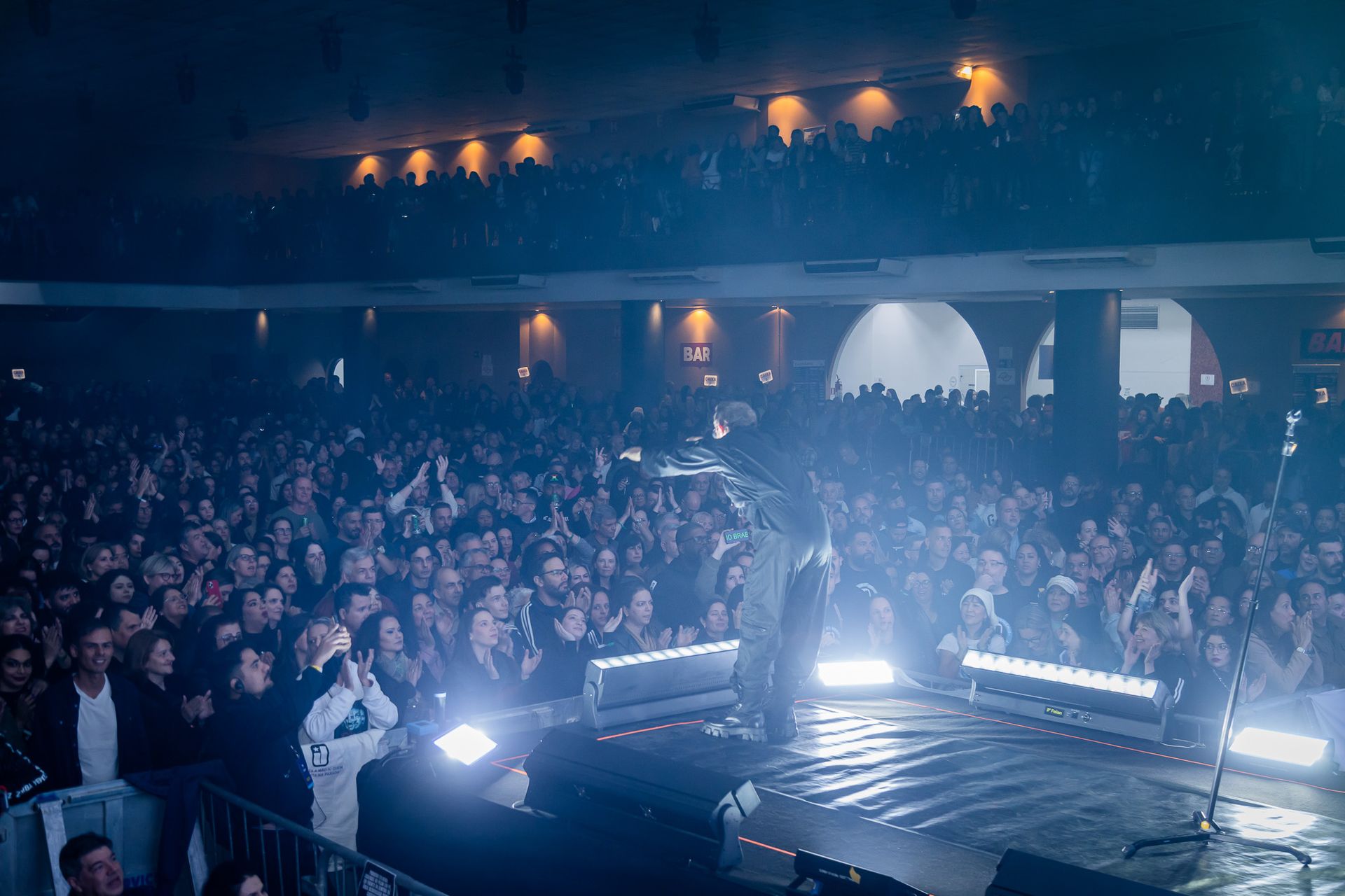 Um artista se apresenta em um palco pouco iluminado diante de uma grande plateia em uma casa de shows fechada.