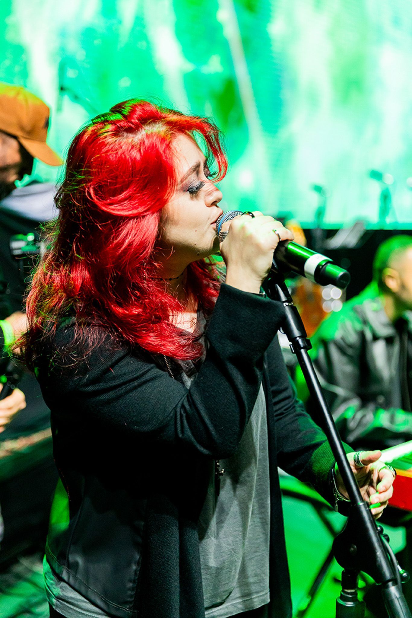 Uma pessoa com cabelos ruivos vibrantes canta em um microfone no palco, iluminada por uma luz verde brilhante.