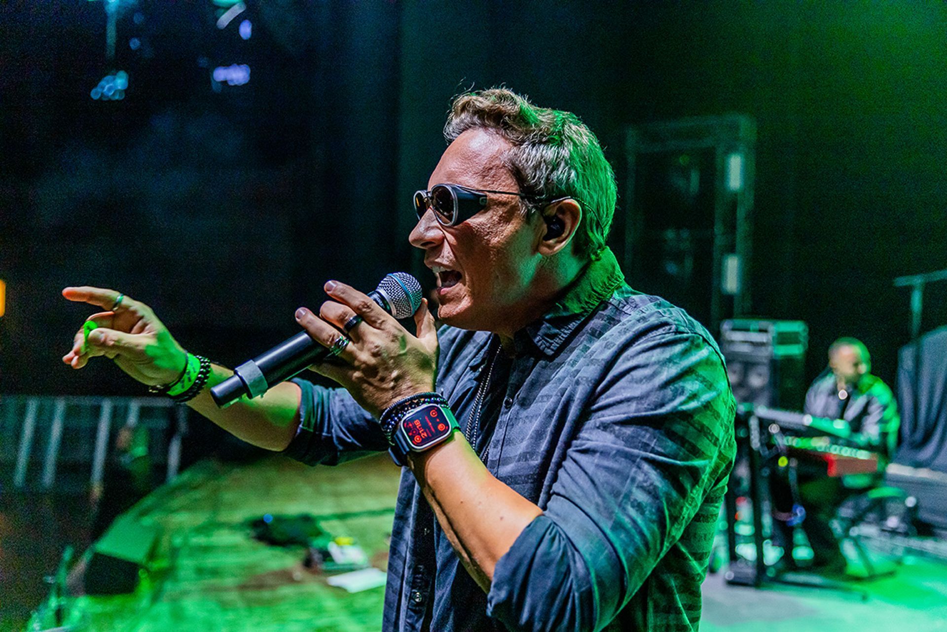 Uma pessoa de óculos escuros e camisa escura canta em um microfone no palco, gesticulando com a mão.