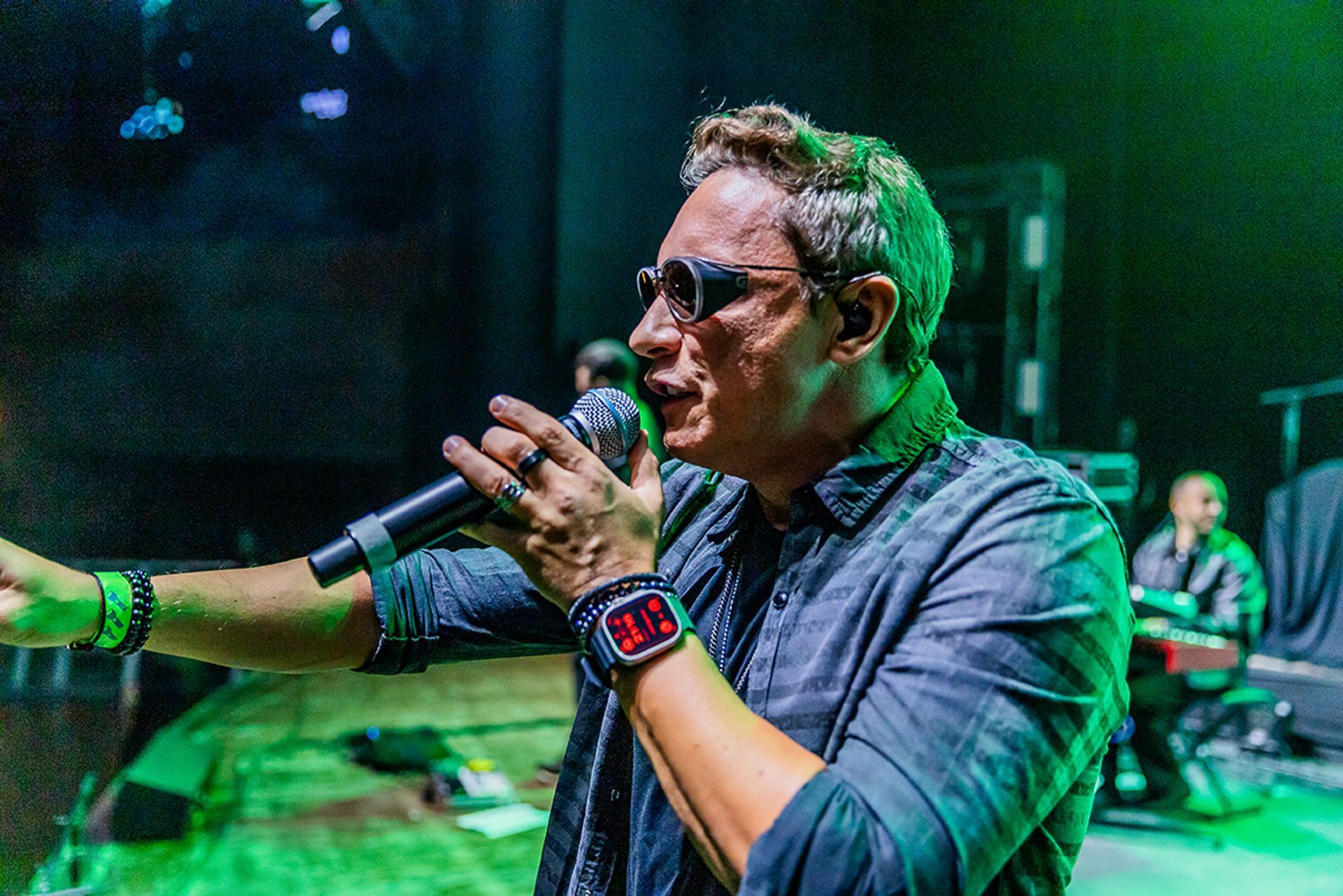 Um cantor se apresentando no palco, segurando um microfone, usando óculos escuros e um relógio digital, sob luz verde.
