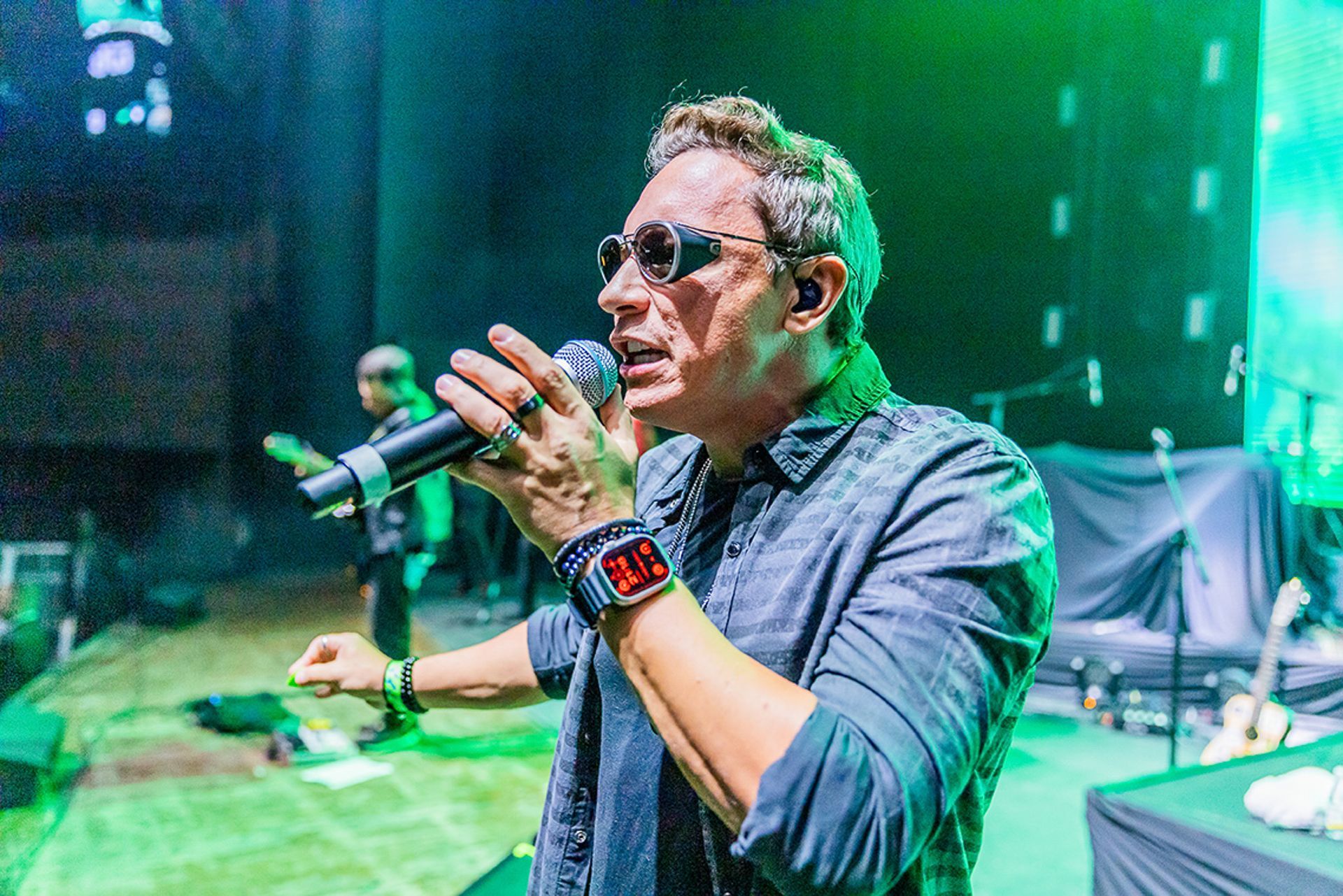 Uma pessoa de óculos segurando um microfone e cantando em um palco de concerto com iluminação verde.