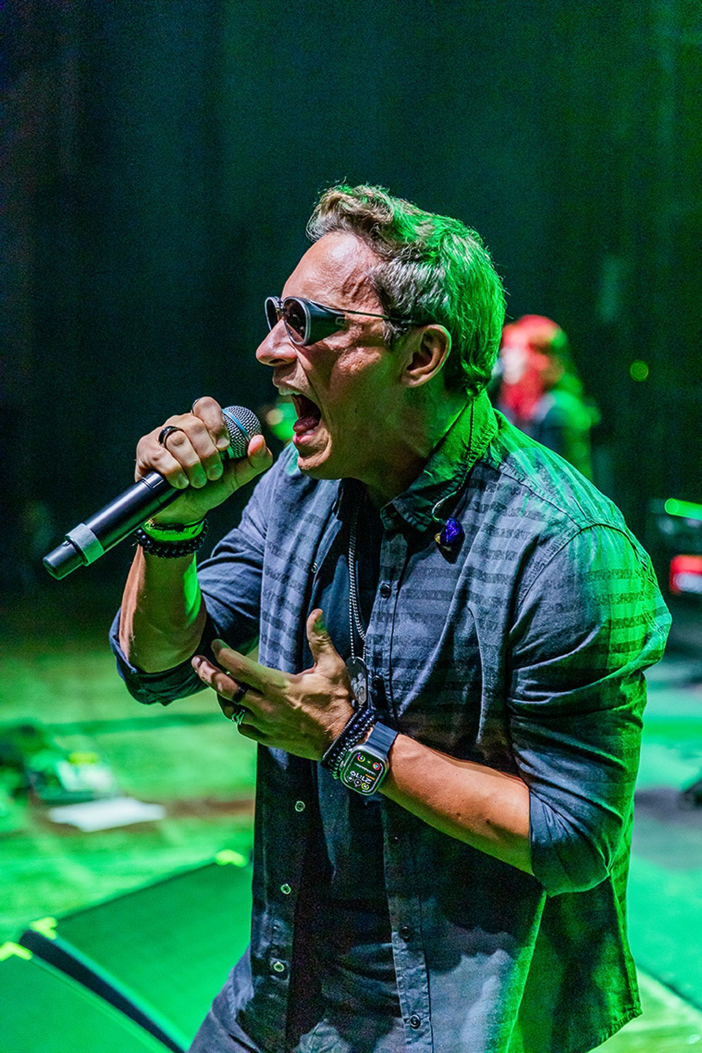 Um cantor de óculos escuros segura um microfone e canta com intensidade em um palco iluminado por luz verde.
