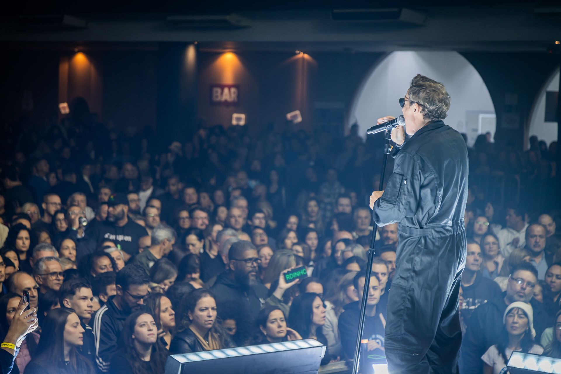 Um artista se apresenta no palco, cantando em um microfone diante de uma grande plateia em um local fechado com pouca luz.