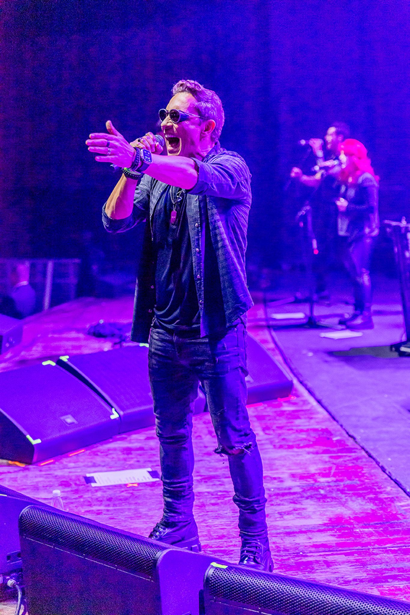 Um artista cantando em um microfone em um palco iluminado por uma luz roxa brilhante, com um membro da banda tocando.