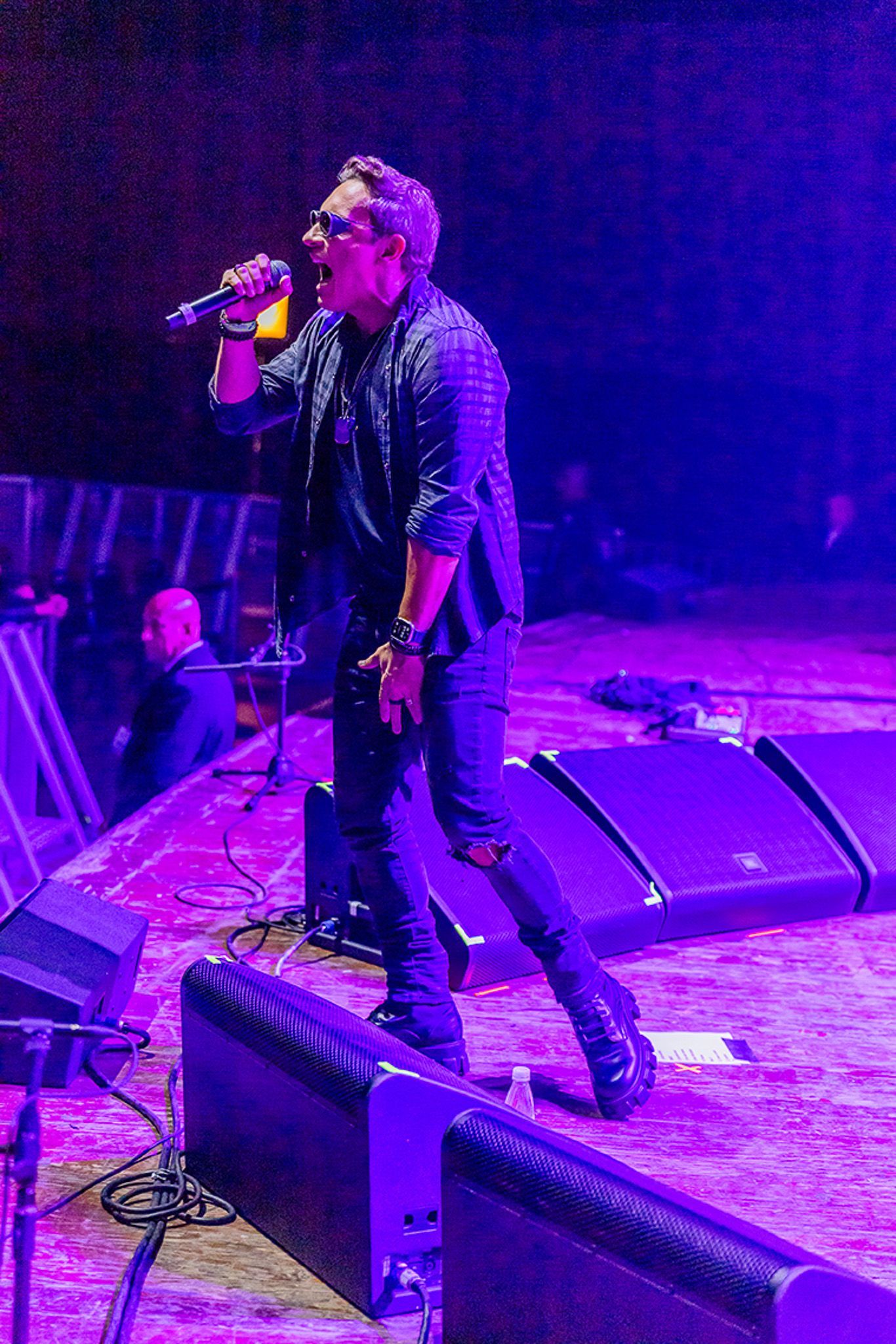 Um vocalista vestido de preto cantando em um microfone em um palco de concerto iluminado por uma intensa luz roxa.