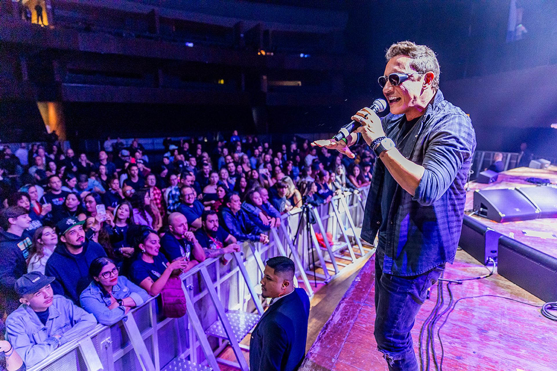 Um artista usando óculos escuros e uma camisa estampada canta em um microfone no palco antes de um grande show lotado.