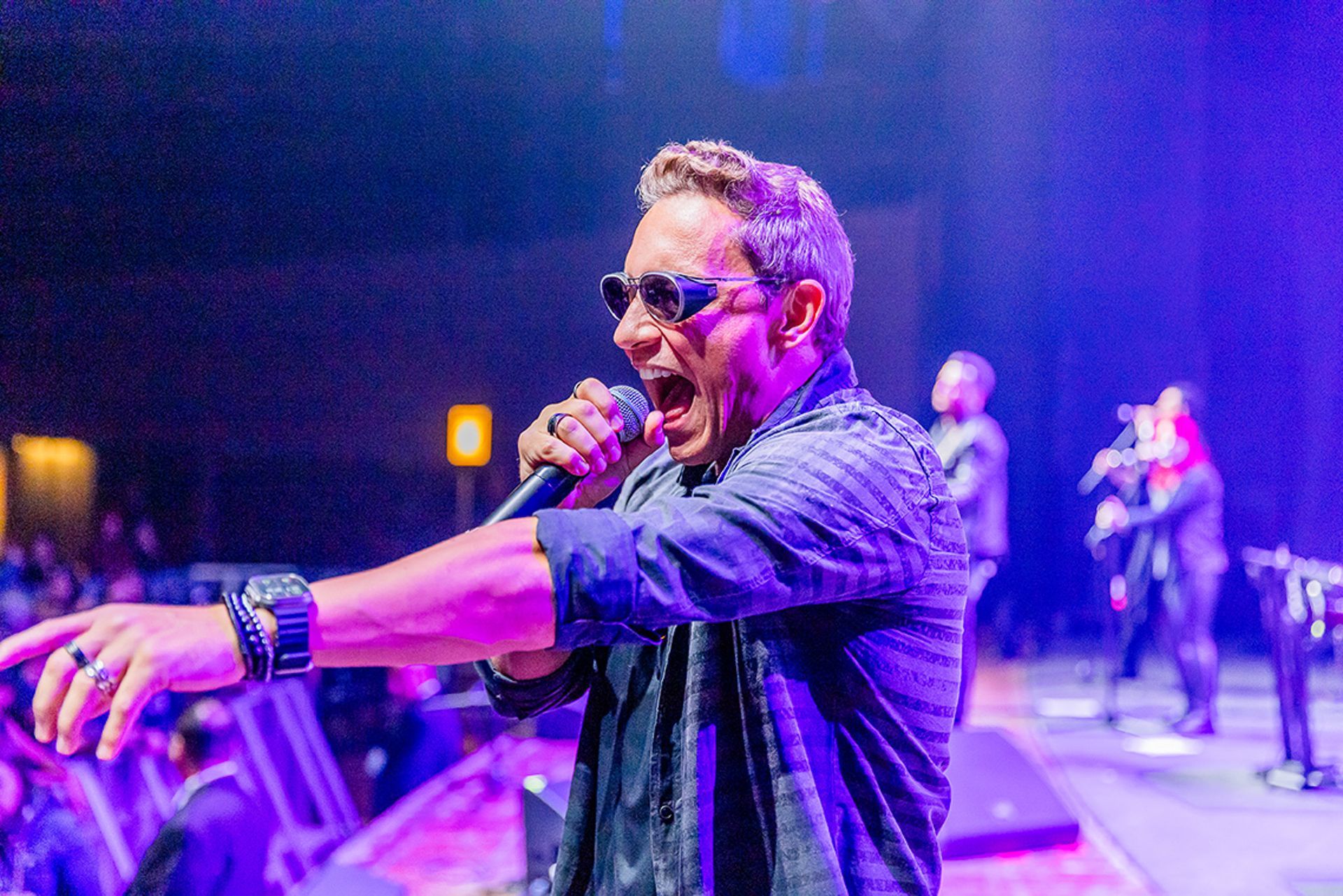 Um artista cantando intensamente em um microfone no palco, apontando para a plateia sob iluminação azul.