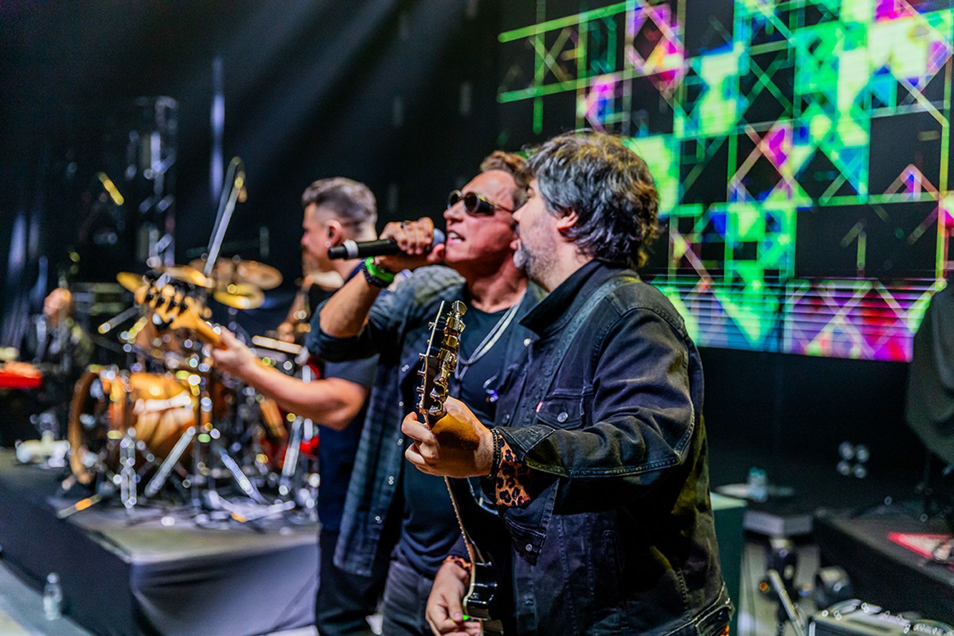 Três músicos se apresentam no palco com instrumentos, incluindo um vocalista de óculos escuros e um guitarrista.