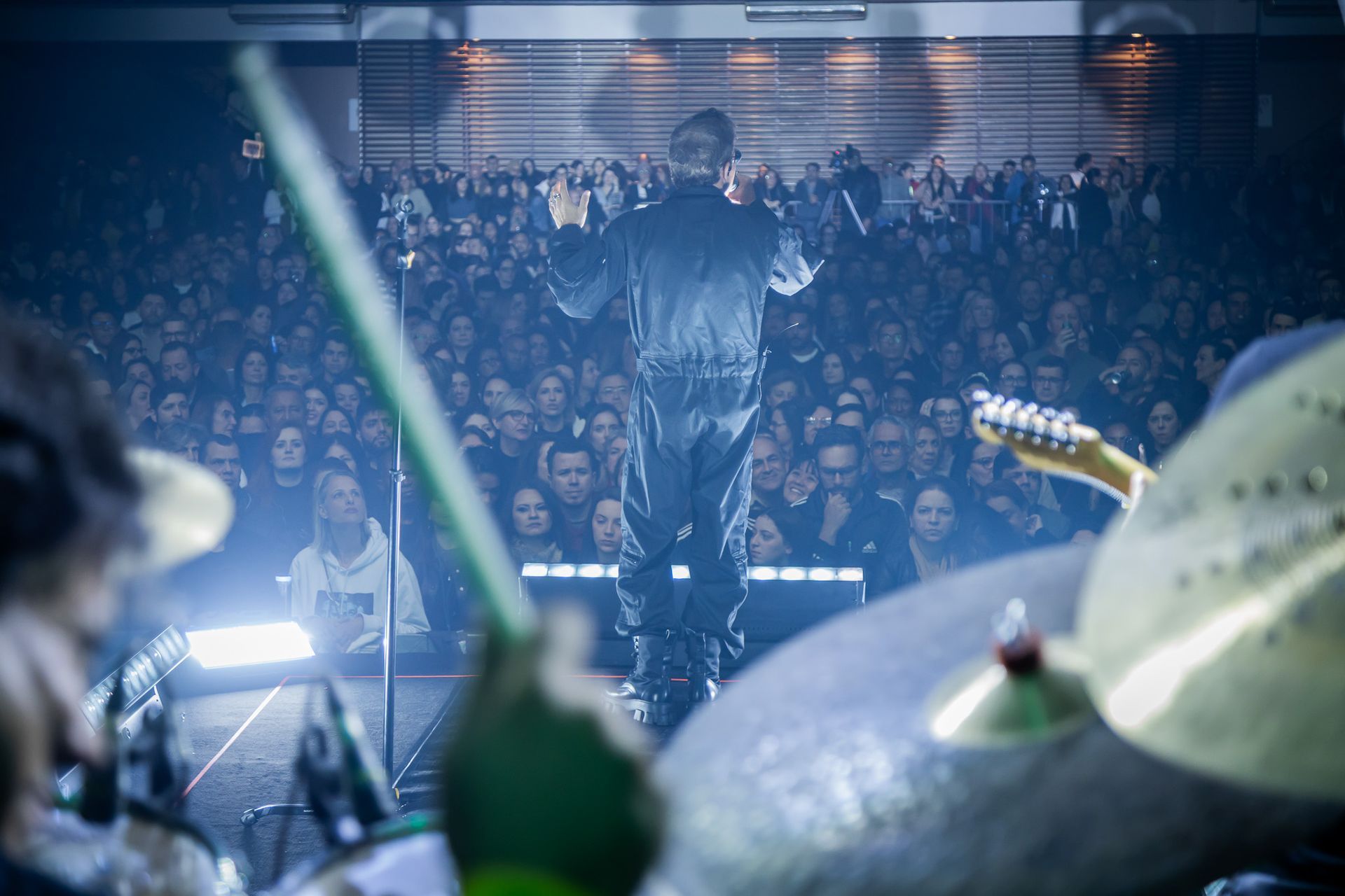 Um artista de terno preto está em um palco, de frente para uma grande plateia iluminada em uma casa de shows.