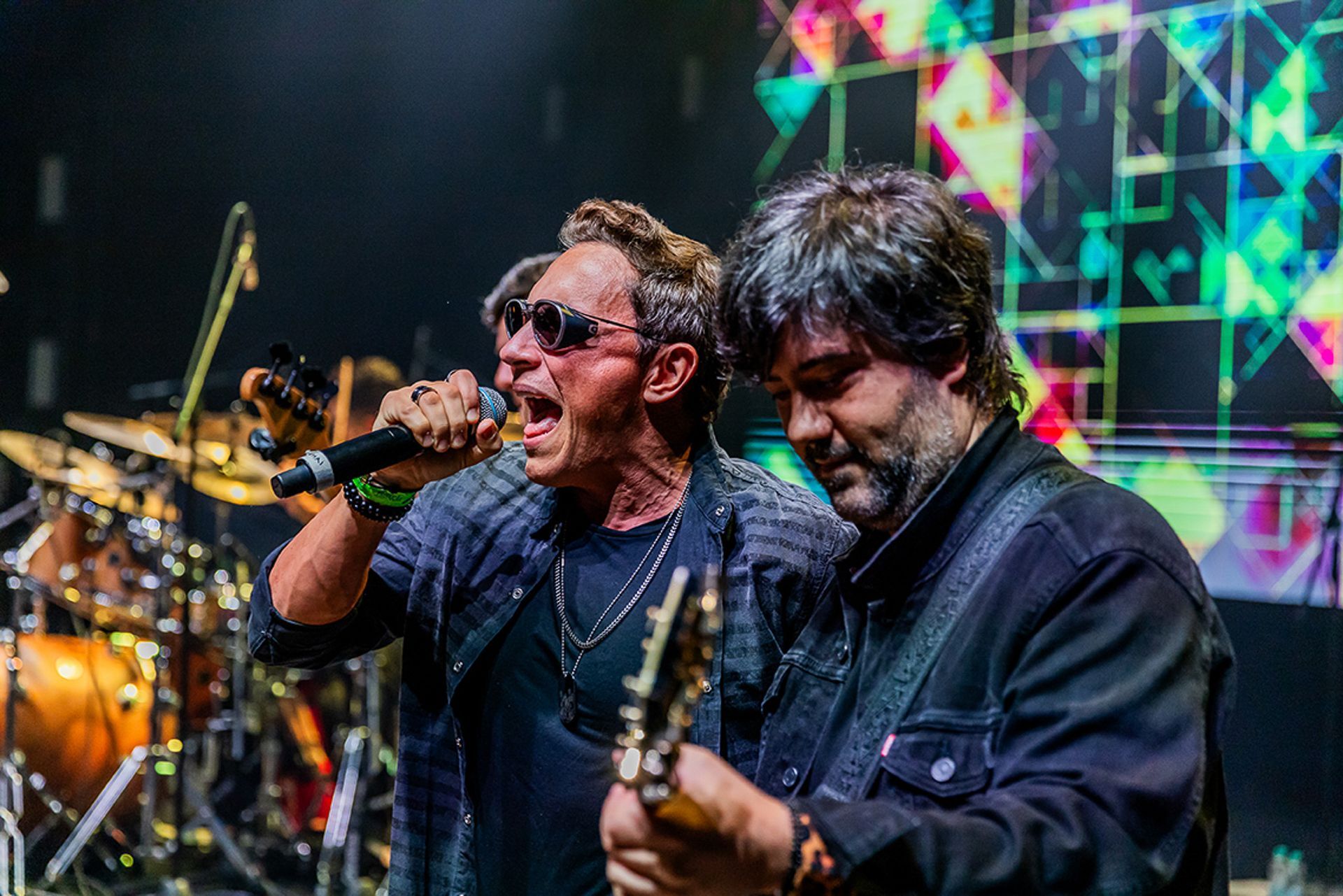 Dois músicos se apresentam no palco; um canta ao microfone usando óculos escuros, o outro toca guitarra elétrica.