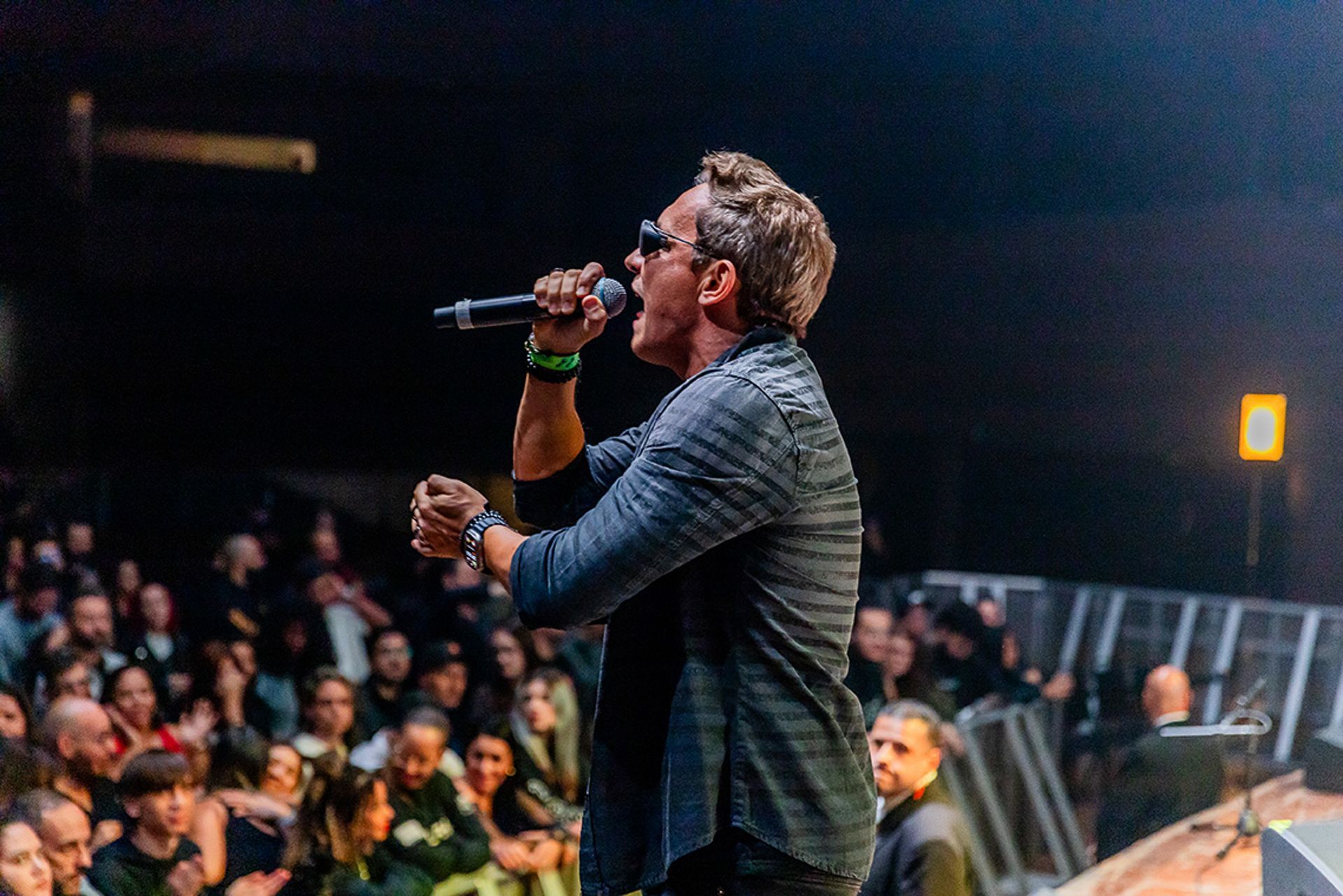 Um artista cantando ao microfone em um palco diante de uma plateia em um local com pouca luz.