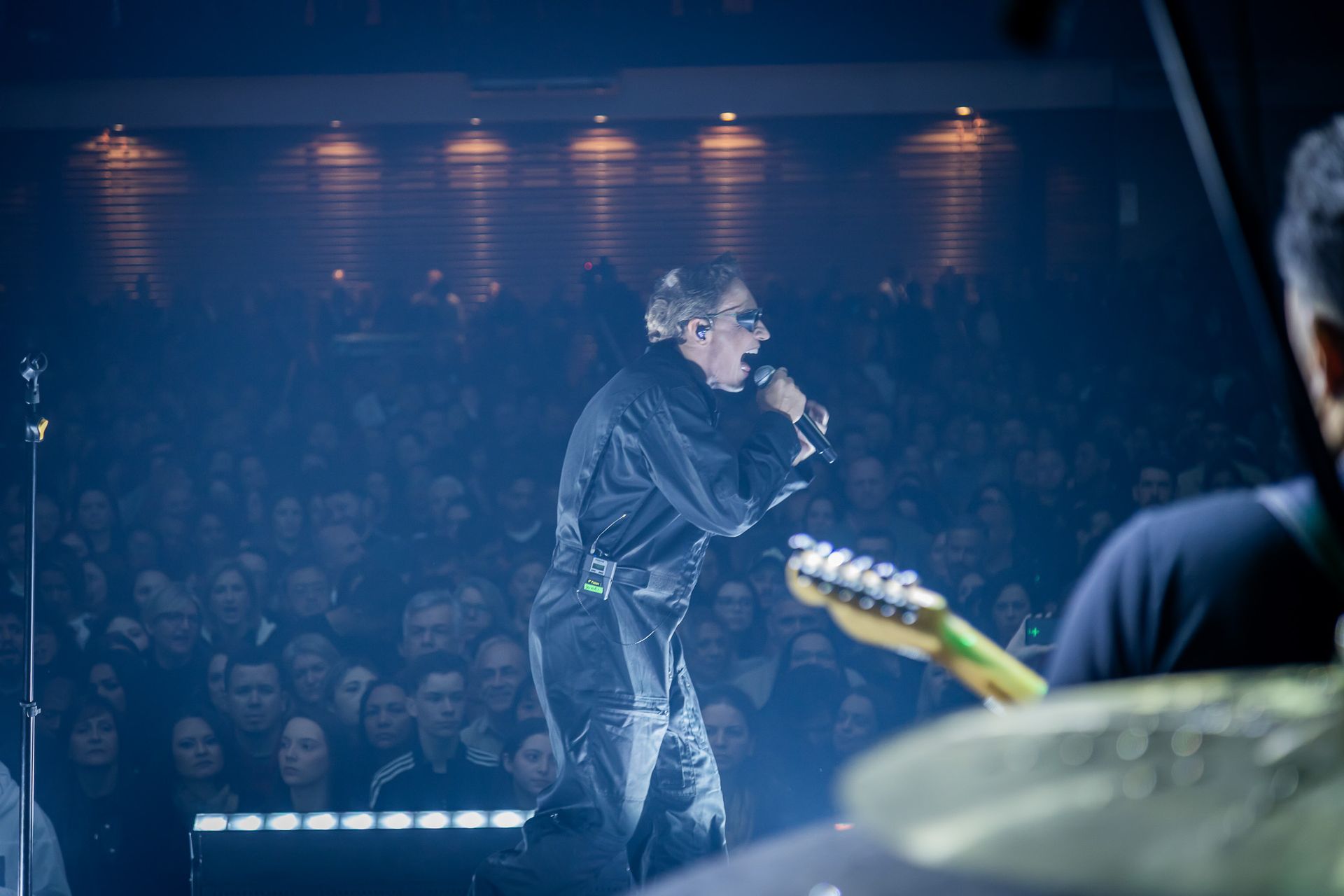 Um artista cantando ao microfone em um palco de concerto, de frente para a plateia sob iluminação cênica azul.
