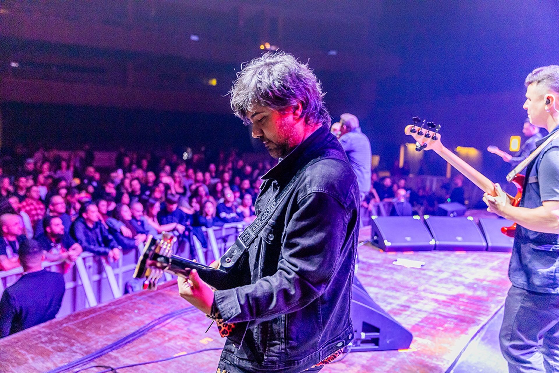 Músicos se apresentam em um palco bem iluminado diante de uma grande plateia em uma casa de shows com pouca luz.
