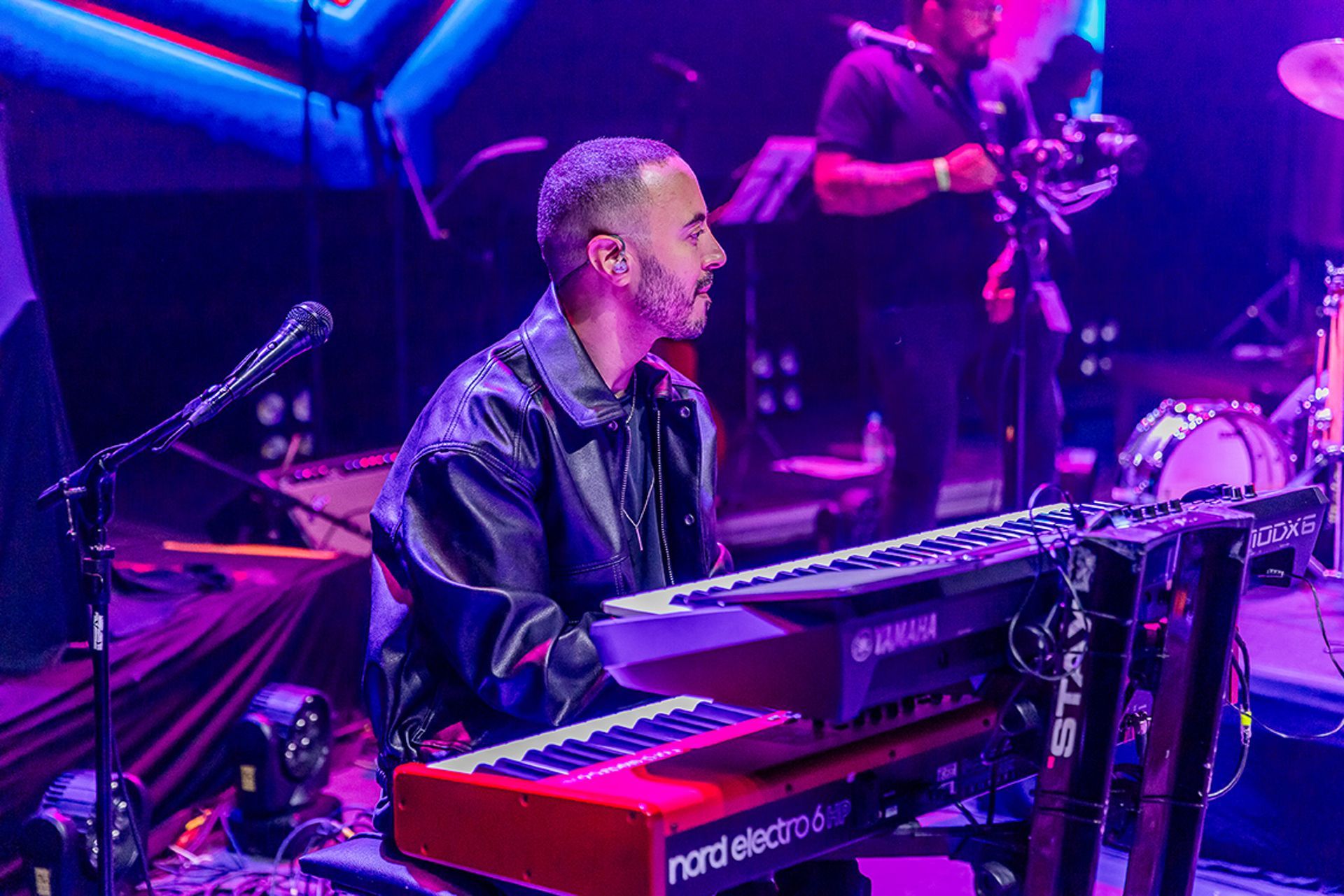 Um músico toca um teclado vermelho no palco, iluminado por luzes roxas, com um pedestal de microfone em primeiro plano.