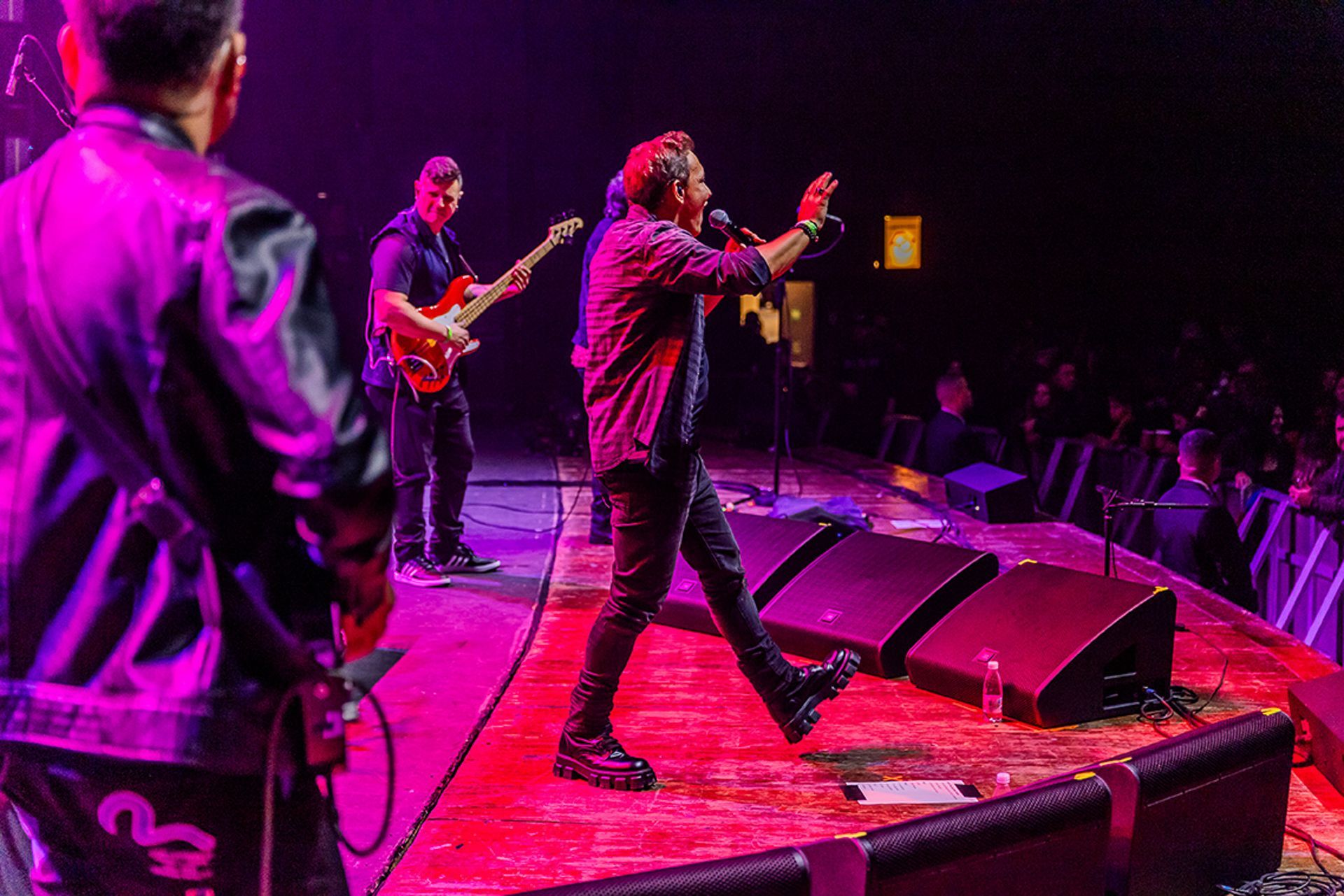 Uma banda se apresenta em um palco com uma iluminação vibrante em tons de roxo e vermelho, com o vocalista gesticulando.