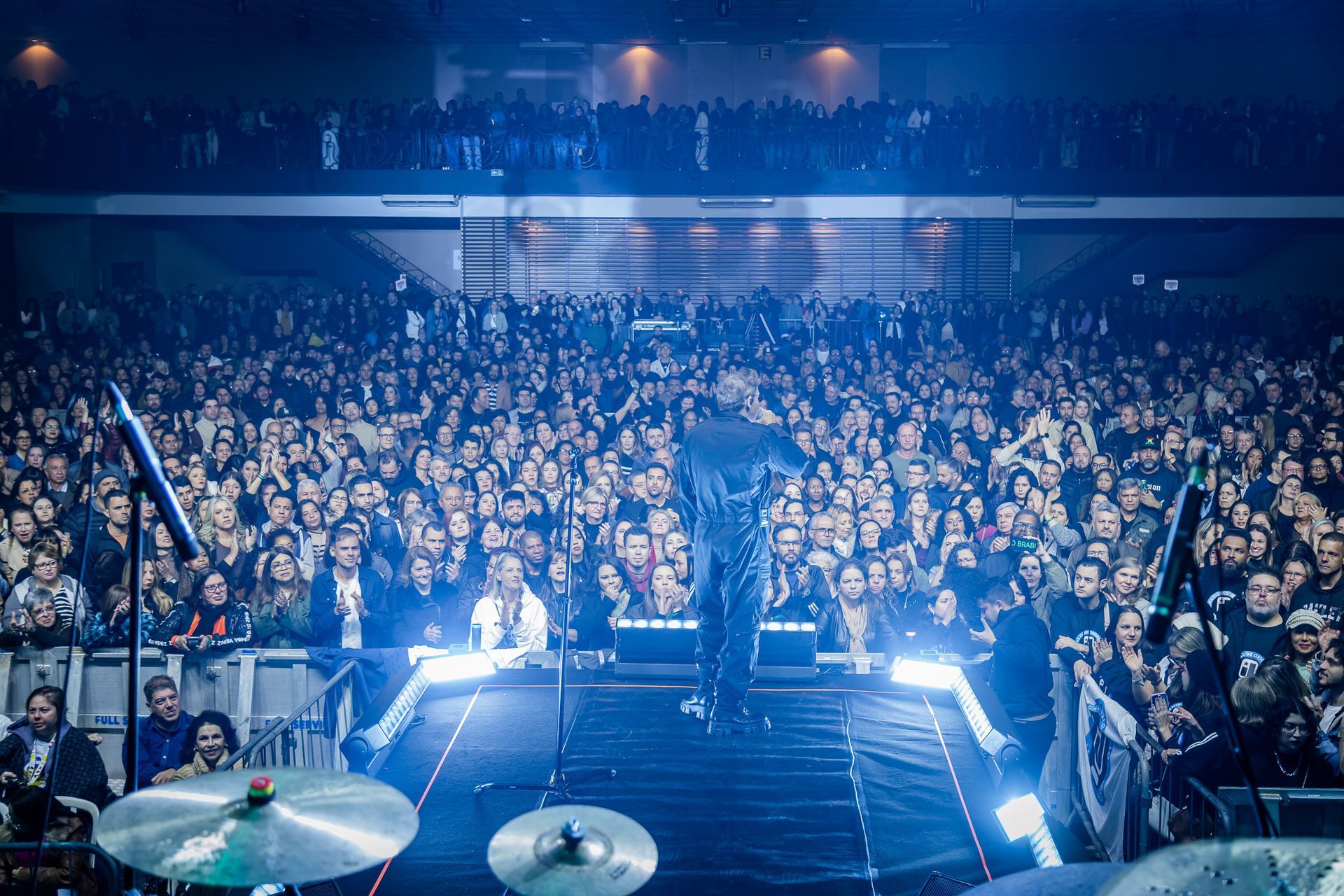 Um artista está em um palco, visto por trás, de frente para uma grande plateia iluminada em uma casa de shows escura.