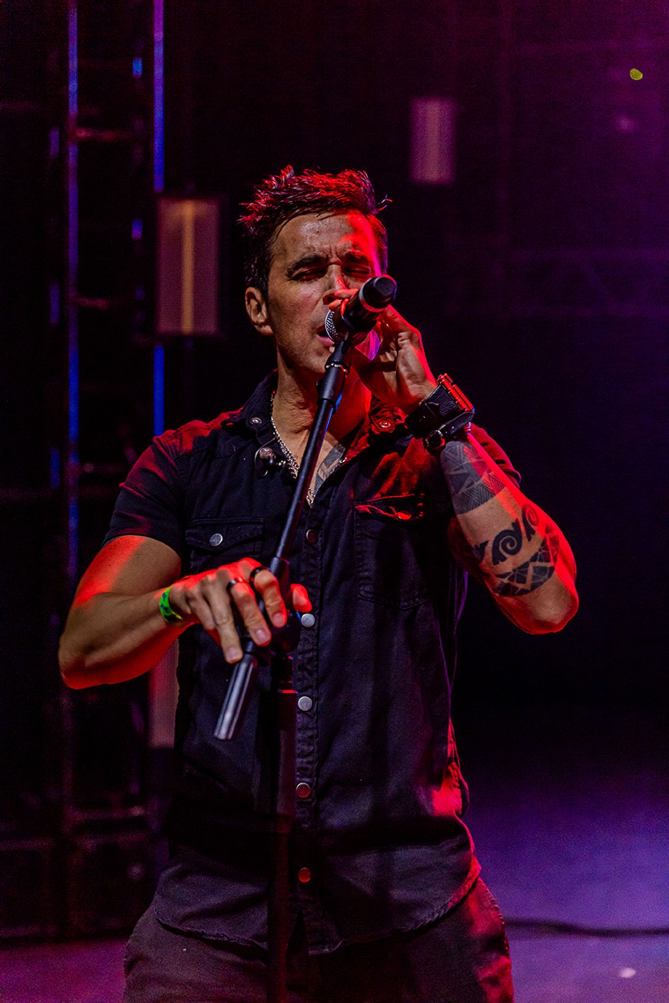 Um artista canta em um palco iluminado com tons de vermelho e roxo, vestindo uma camisa preta e com tatuagens no braço.