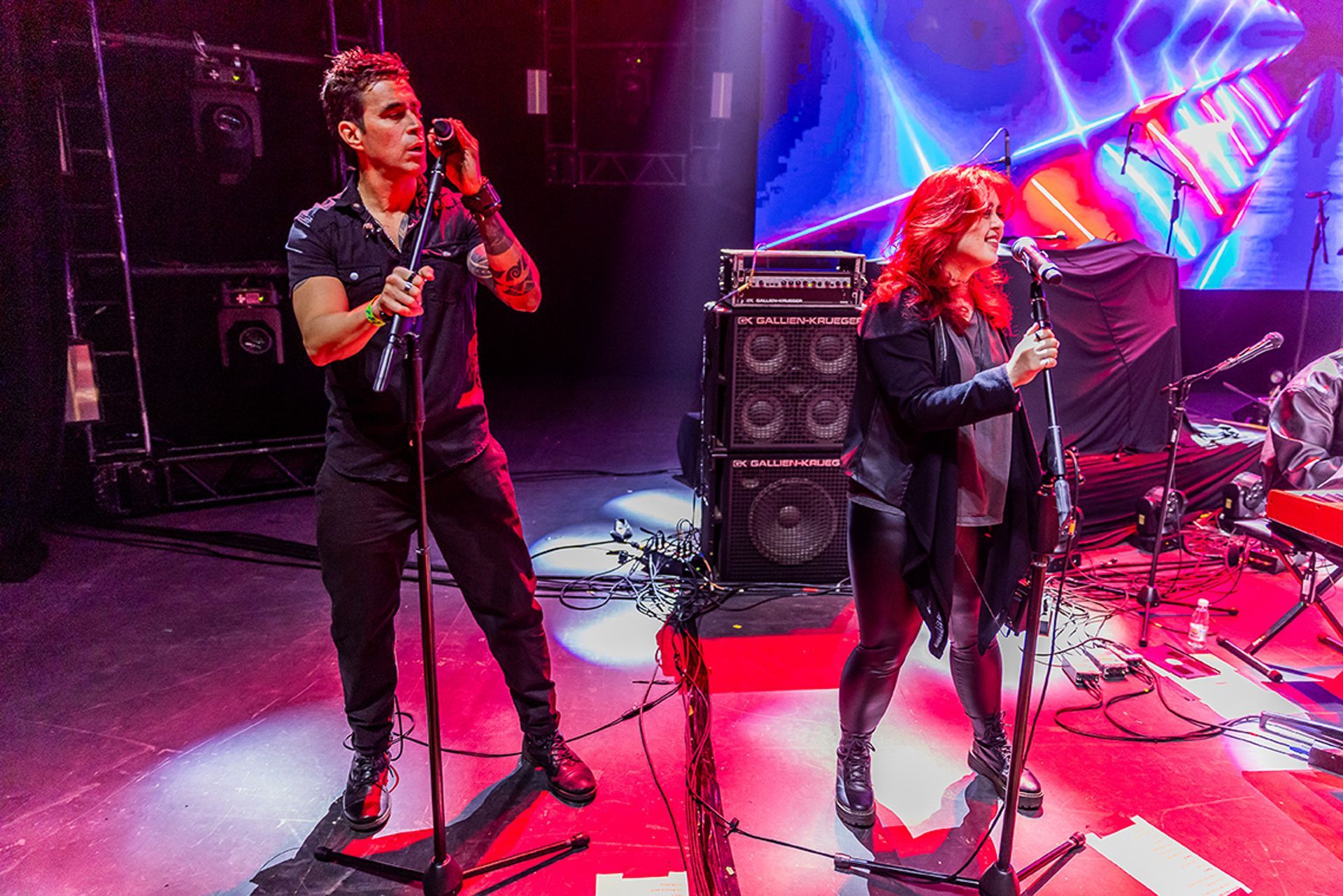 Um músico e uma musicista se apresentam em um palco com um telão digital brilhante ao fundo e grandes caixas de som.