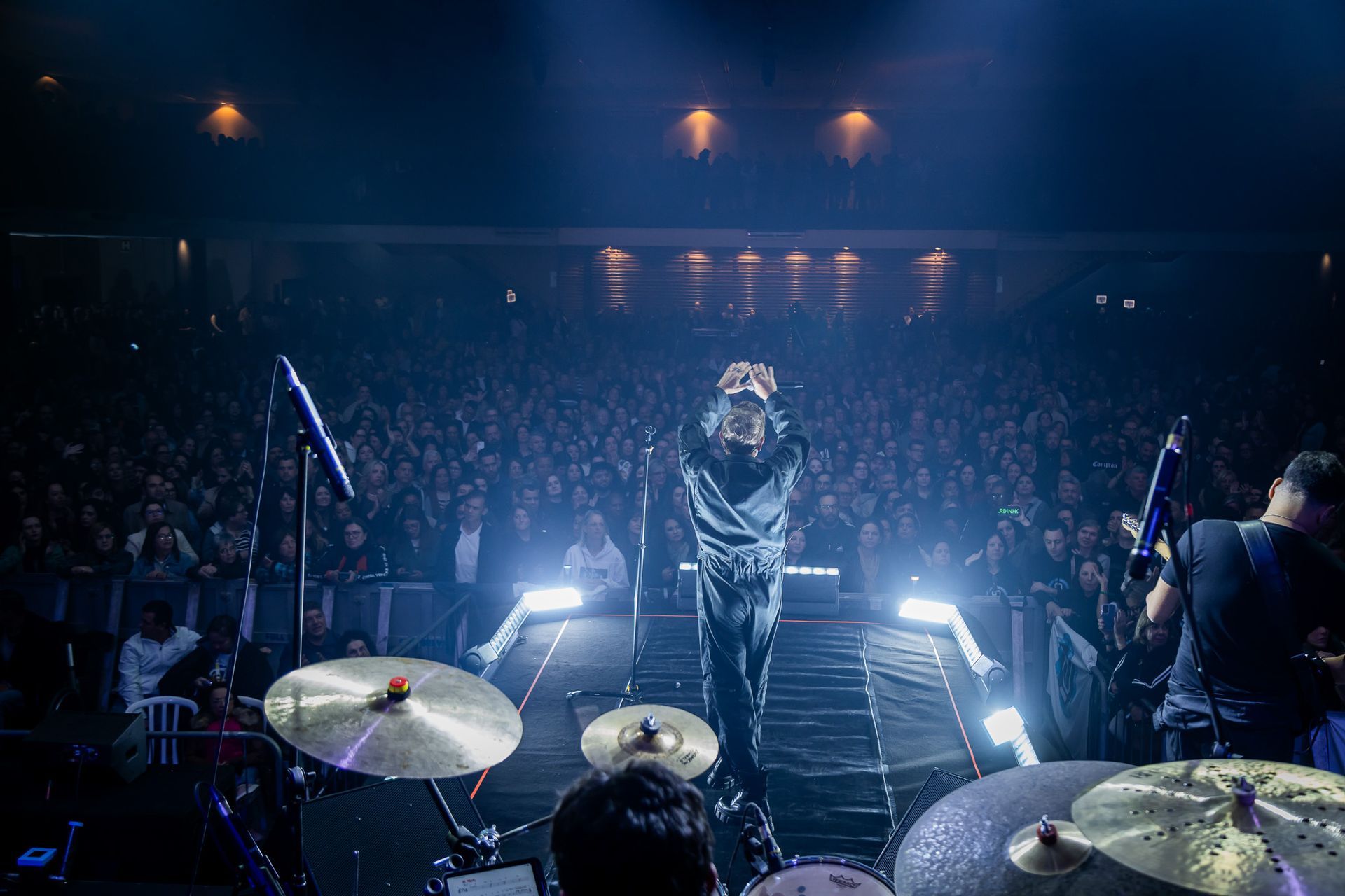 Um artista está de pé no palco de um concerto, de costas para a câmera, de frente para uma grande plateia iluminada.